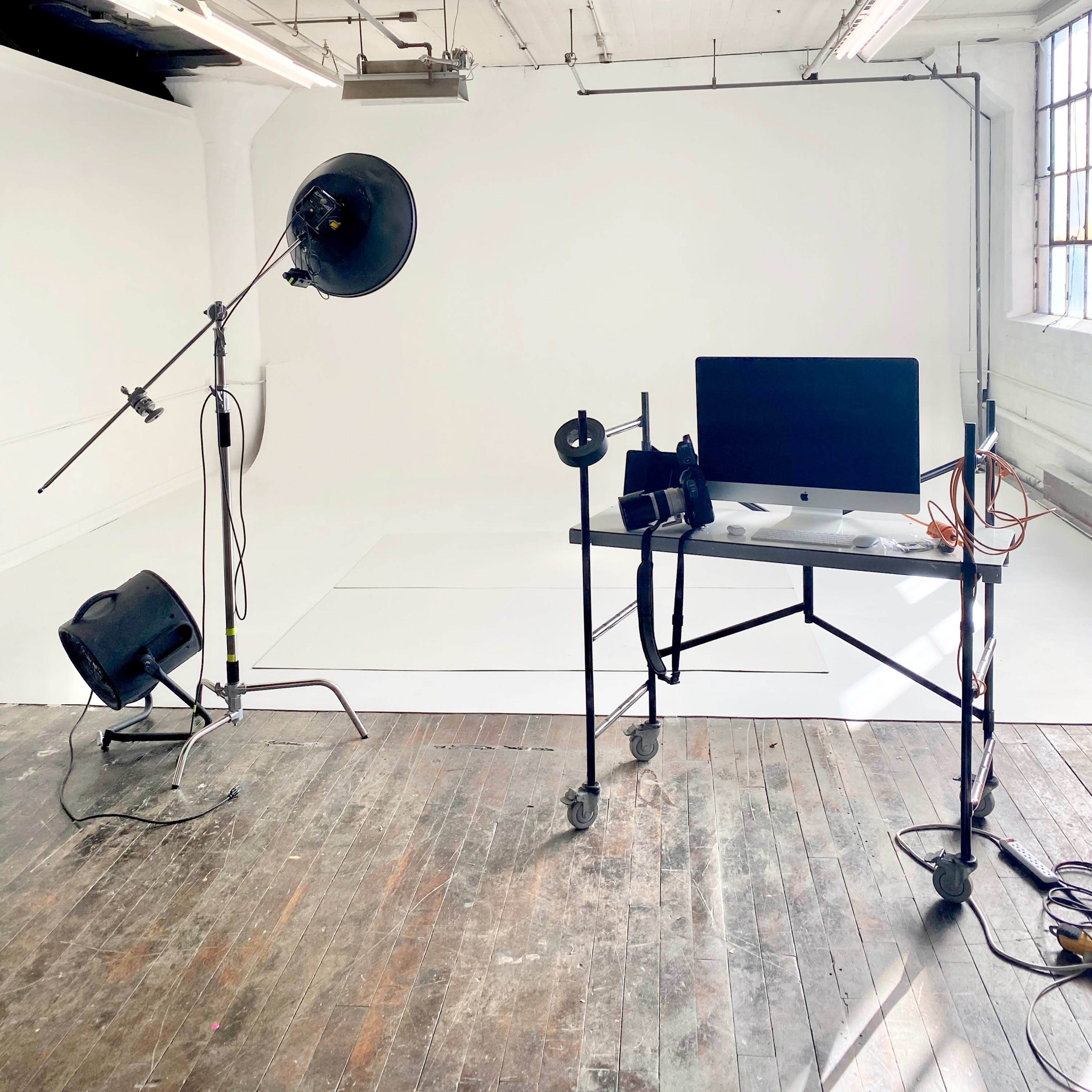 A photography studio with a blank white backdrop, a light stand, and a desk featuring a computer on wheels.