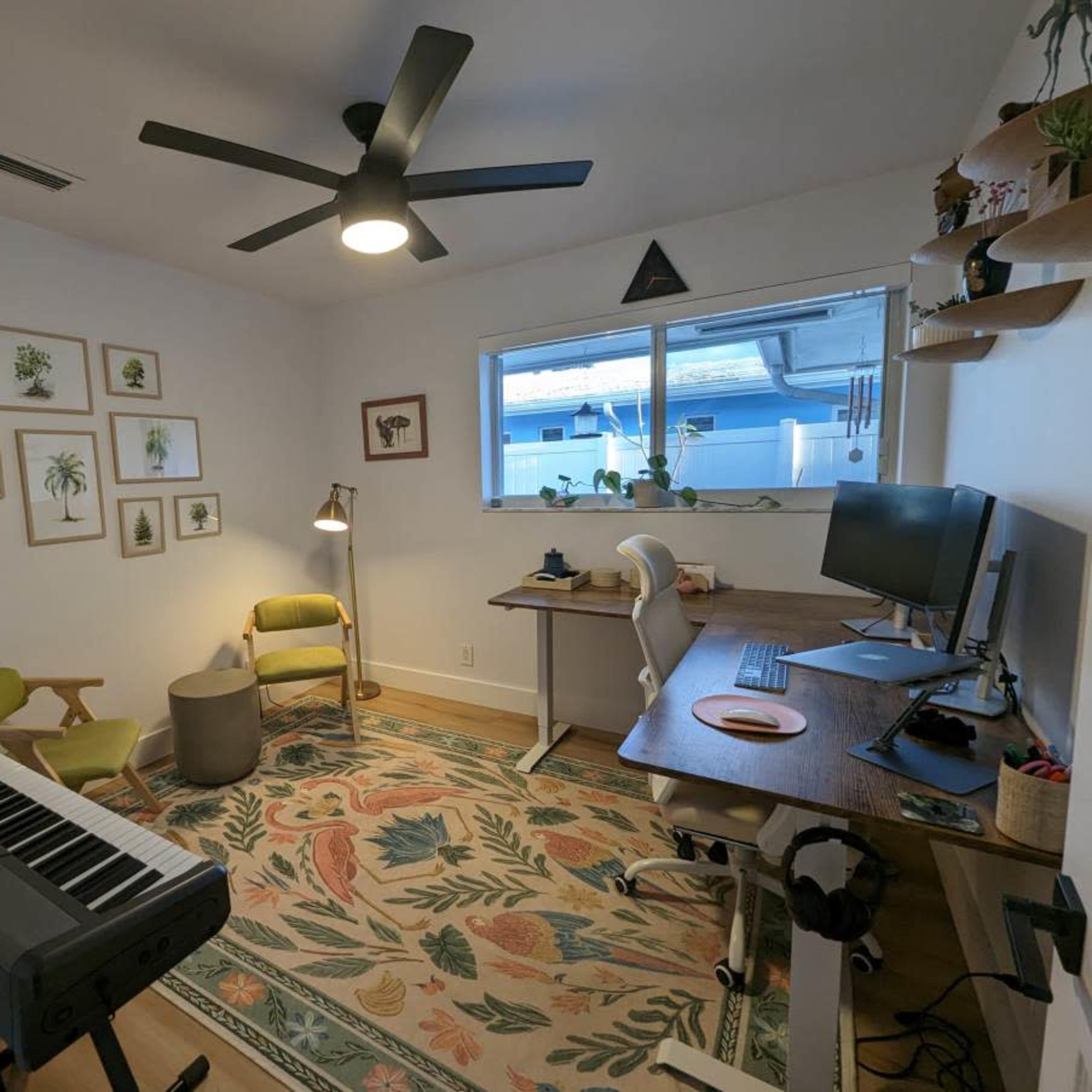A well-organized home office features a keyboard, a desk with a computer, two chairs, and wall art surrounded by plants.