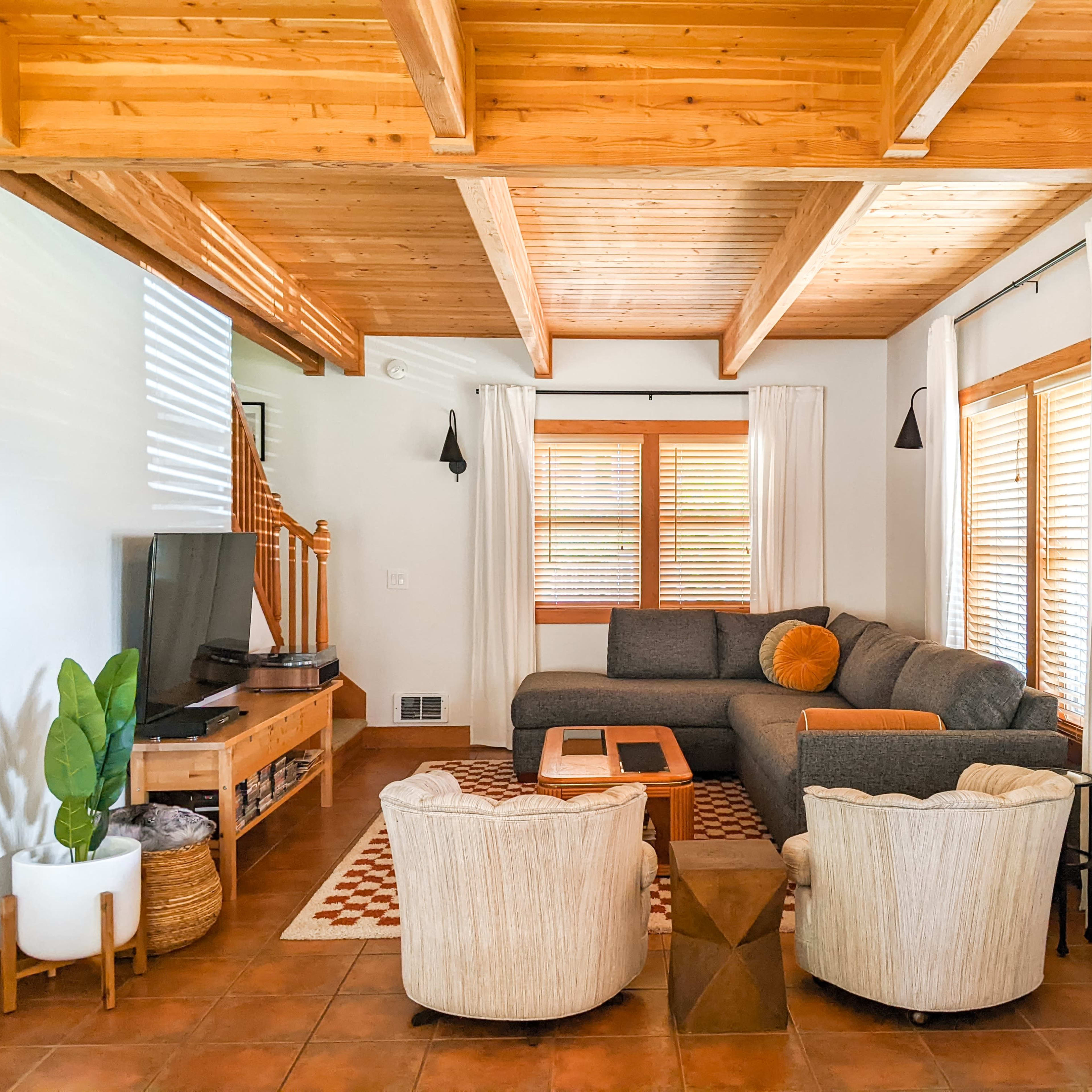 The living room features a sectional sofa, two armchairs, a coffee table, and a television, all arranged under wooden beams with large windows providing natural light.