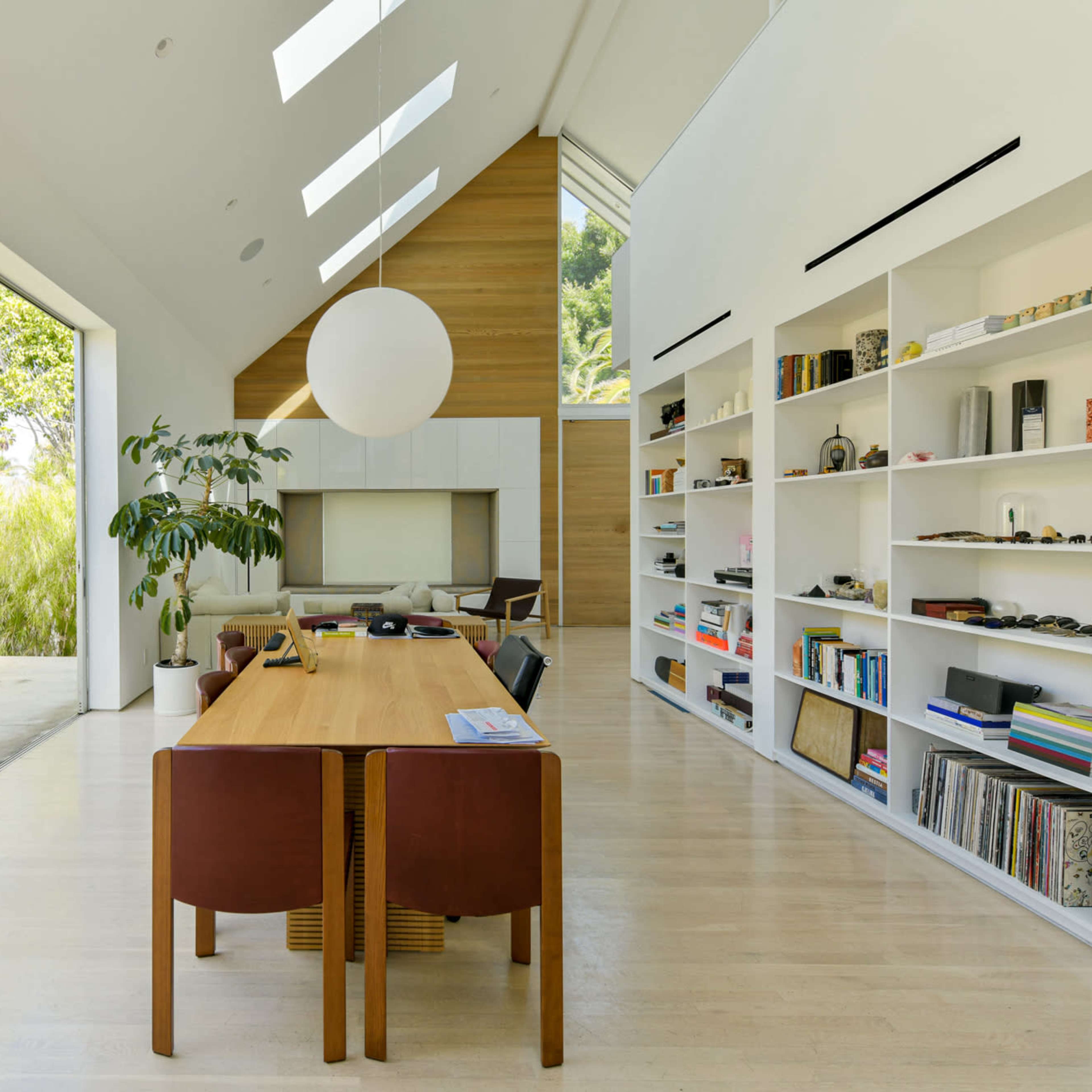 A spacious modern living area features a dining table surrounded by chairs, large windows, and built-in bookshelves.