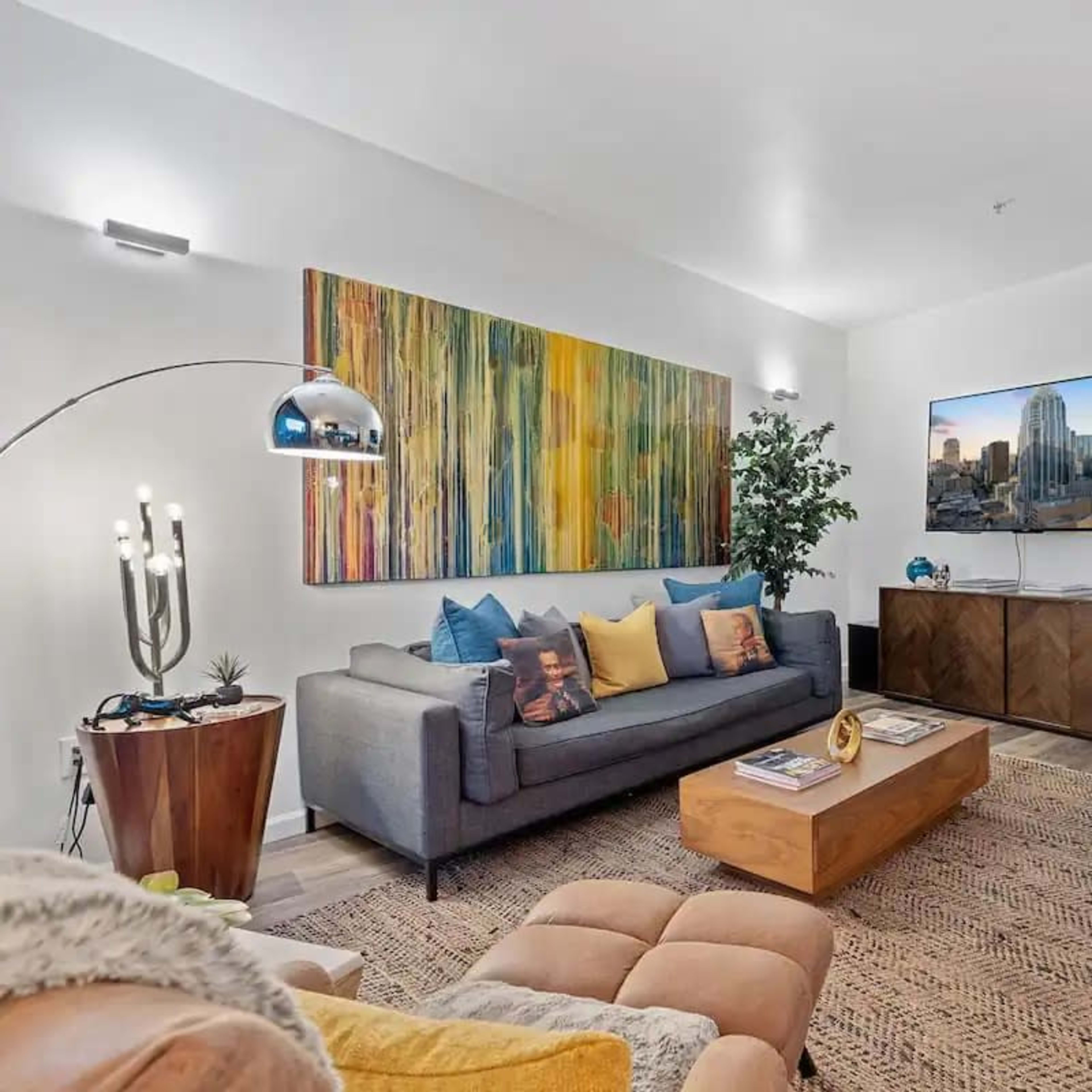 A modern living room featuring a gray sofa with colorful pillows, a wooden coffee table, and a television mounted on a white wall, complemented by vibrant artwork.