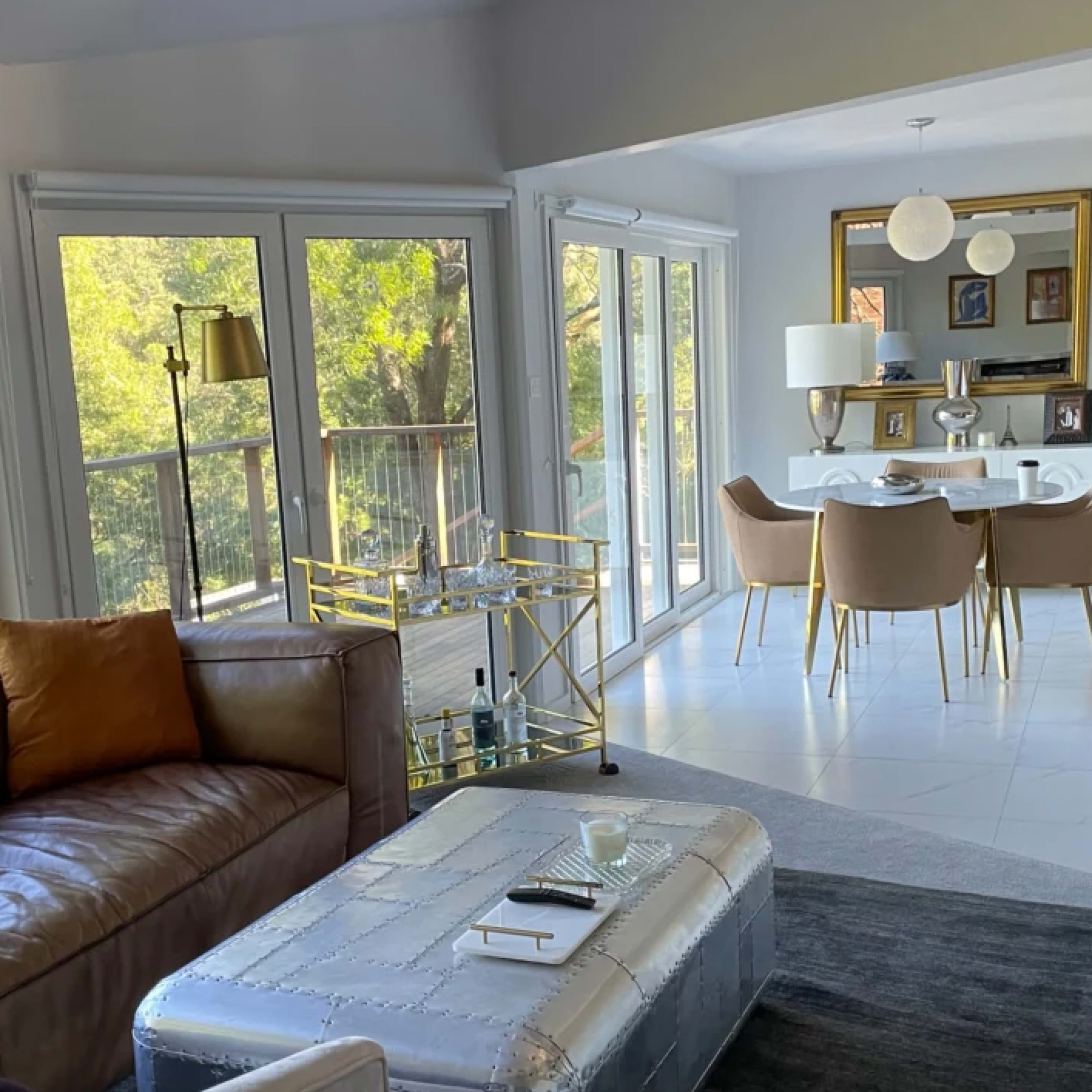 A spacious living area features a brown leather sofa with an orange pillow, a glass coffee table, and a dining space with a white table and beige chairs, all set against large windows overlooking greenery.