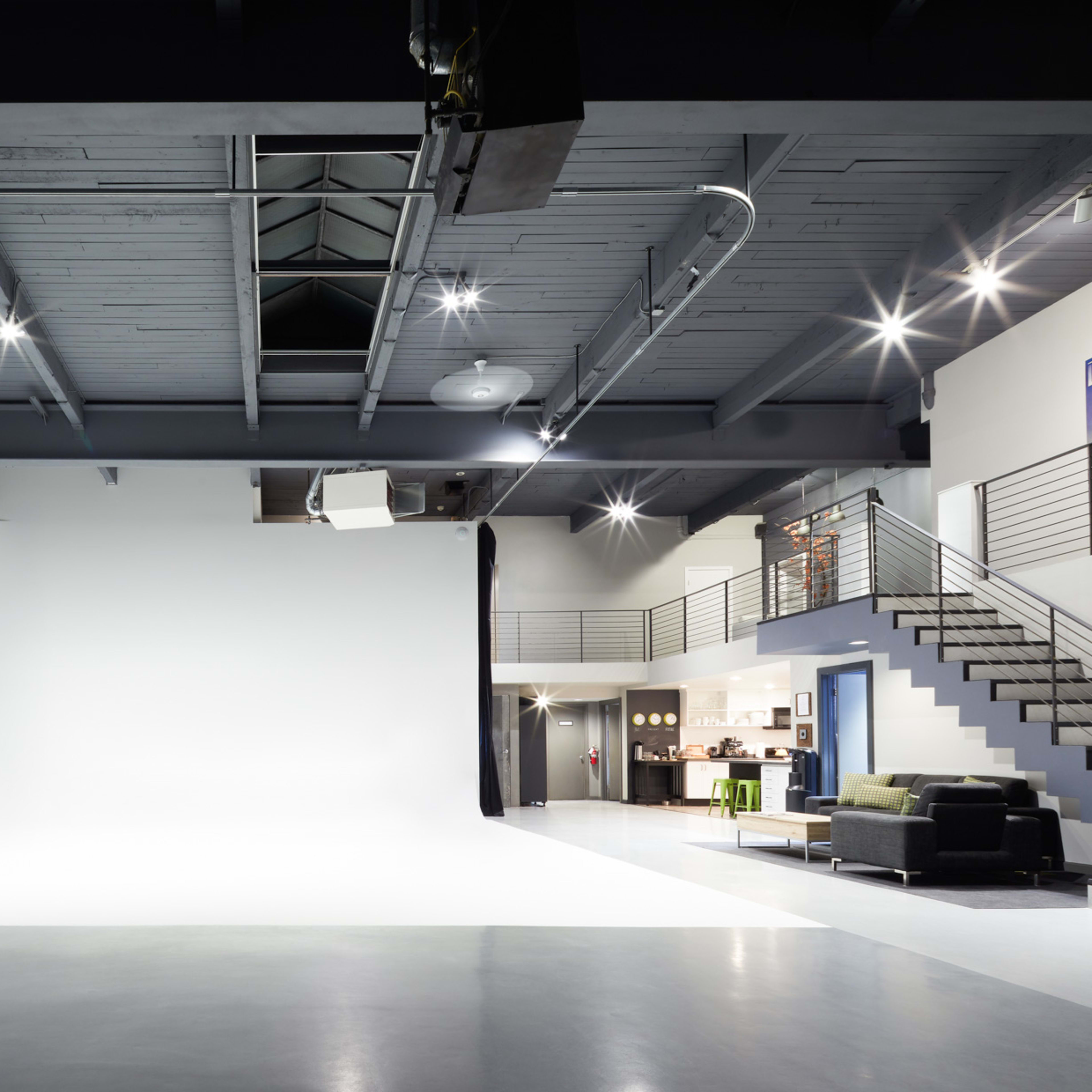 A spacious photo studio with a large white backdrop, polished concrete flooring, and a staircase leading to a second level.