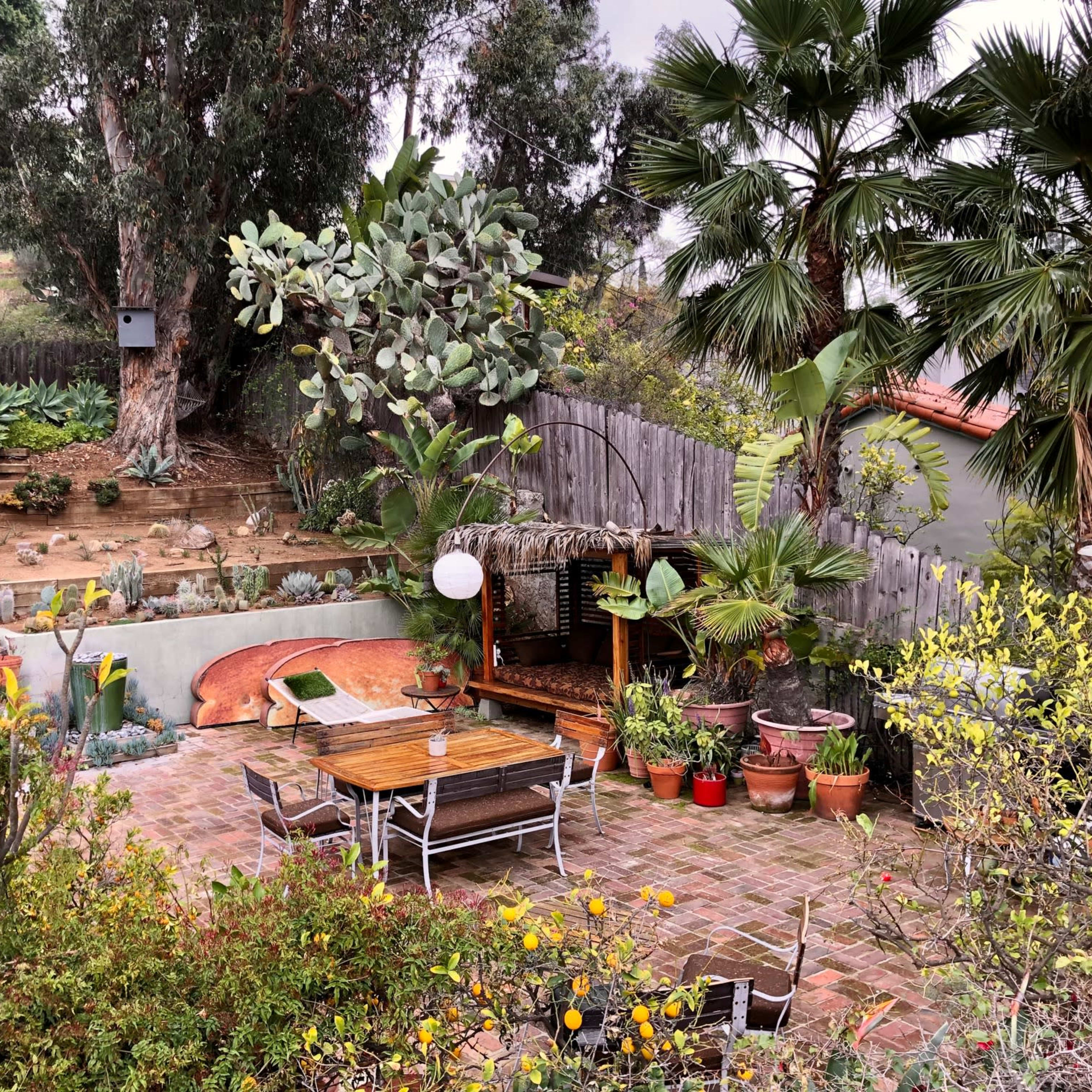 A landscaped garden with a stone patio, wooden dining set, various plants, and a shaded seating area under a pergola.