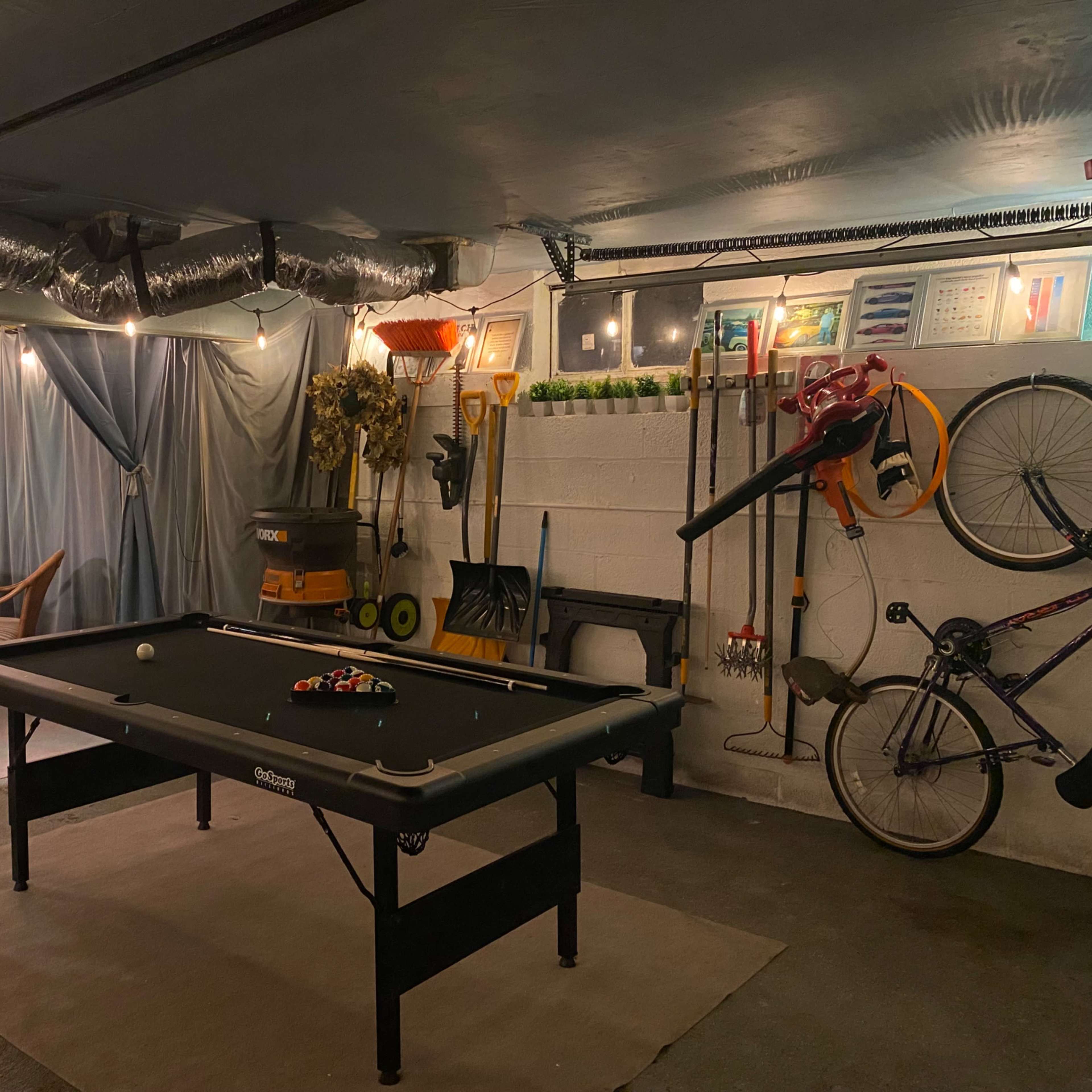 A dimly lit basement recreation area with a pool table, a small dining table, and various tools and bicycles stored on the walls.