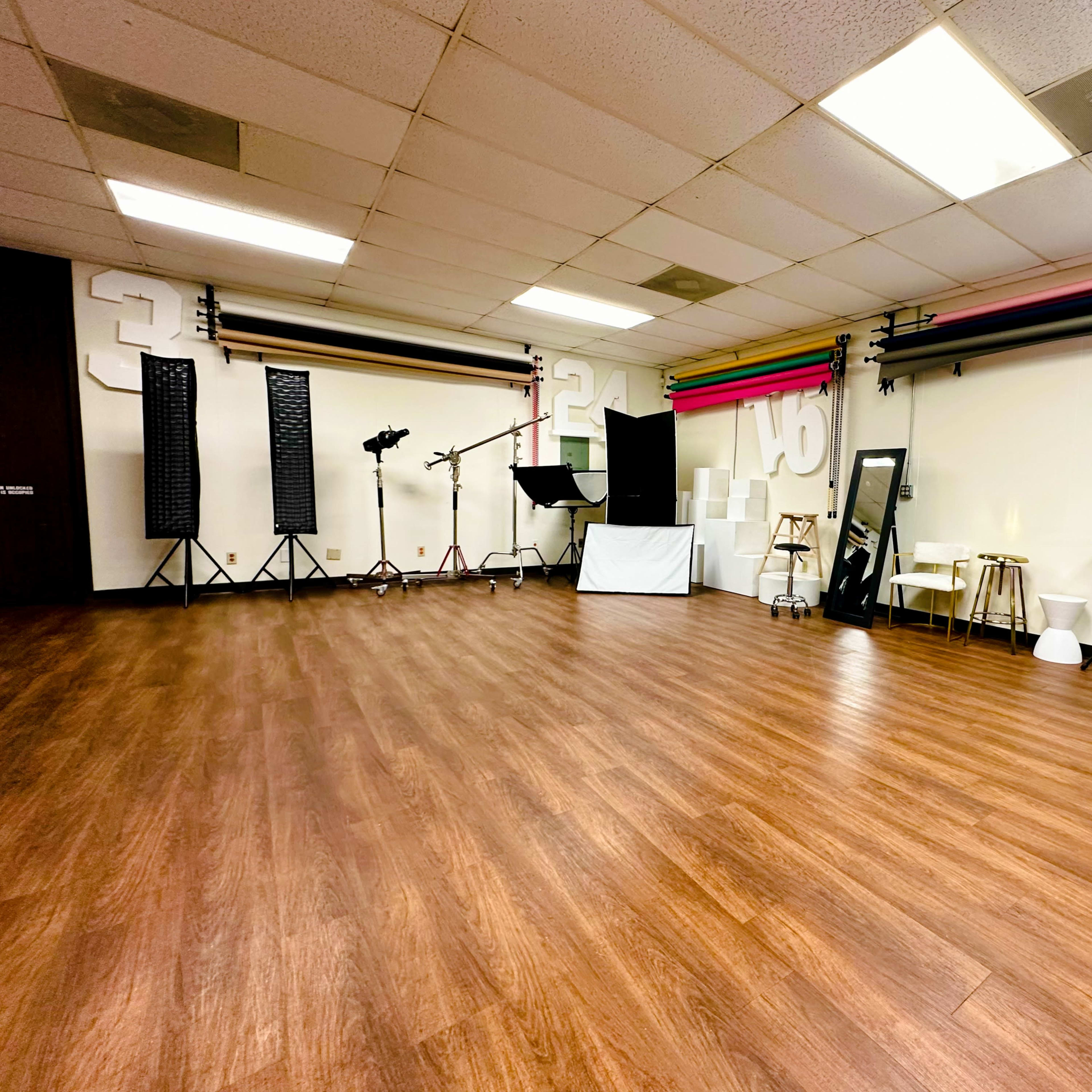 A spacious studio with wooden flooring, equipped with various backdrop rolls, lighting stands, and a mirror.