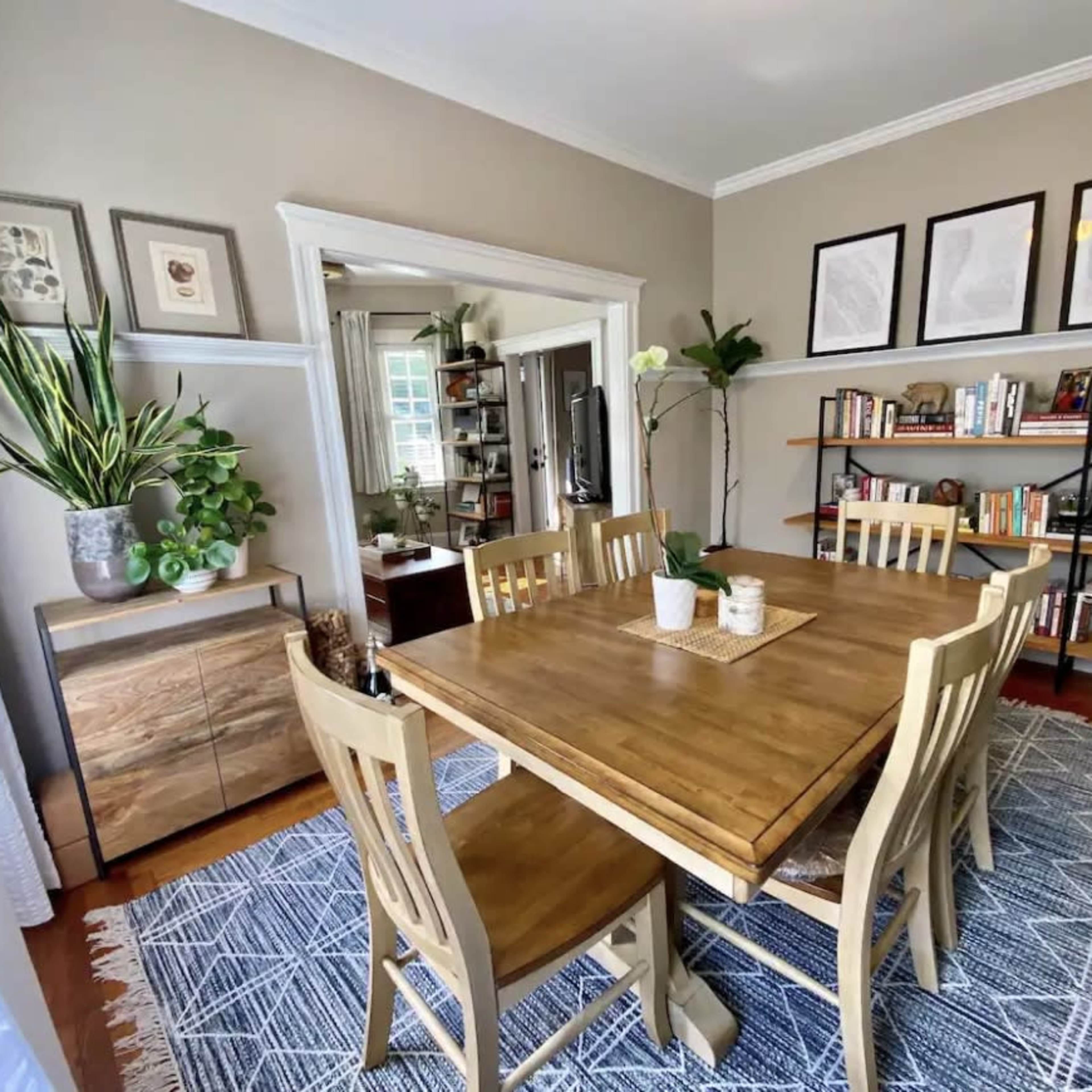 A dining room features a wooden table surrounded by four chairs, with plants and bookshelves in the background, and framed pictures on the wall.