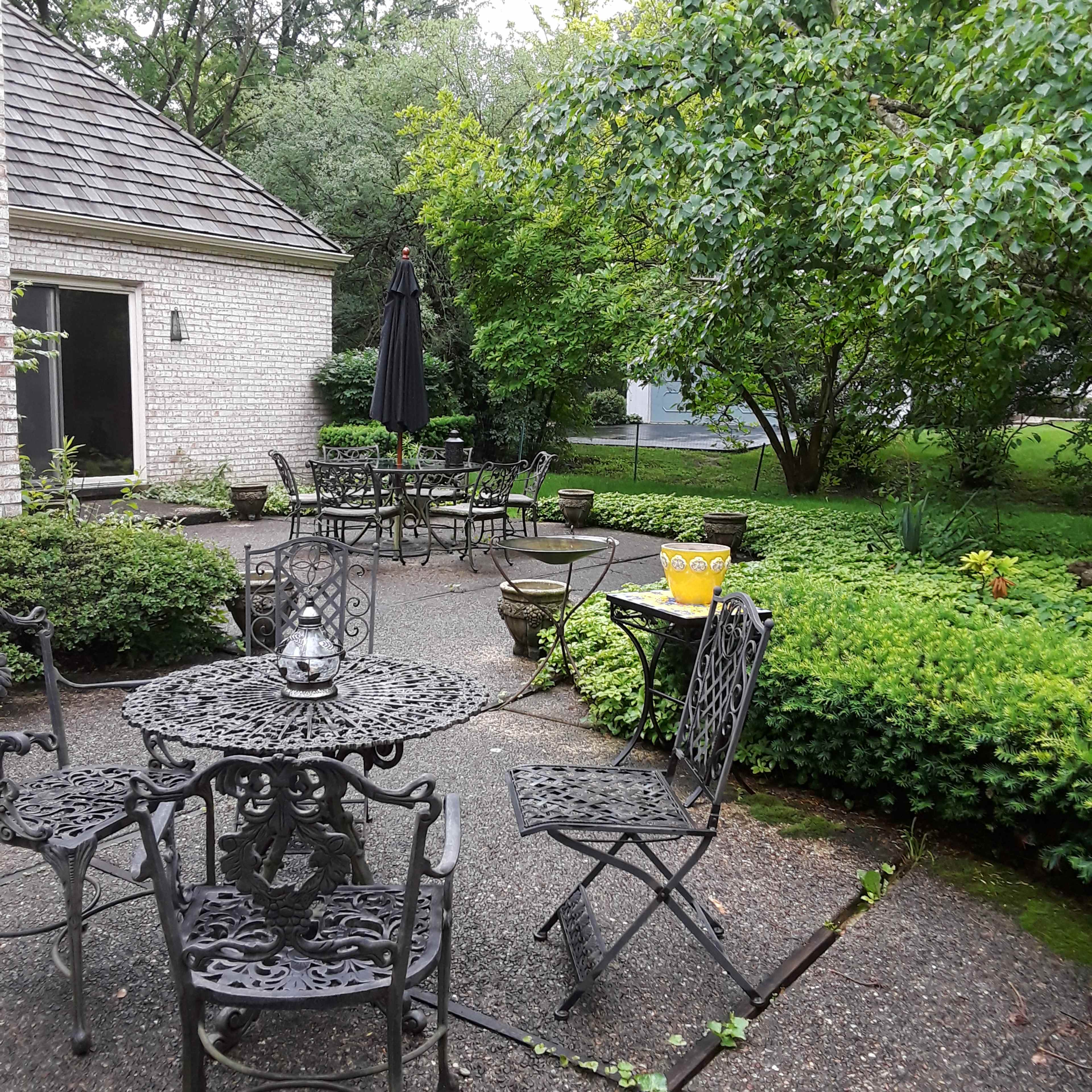 A courtyard features metal tables and chairs surrounded by lush greenery and a stone pathway.