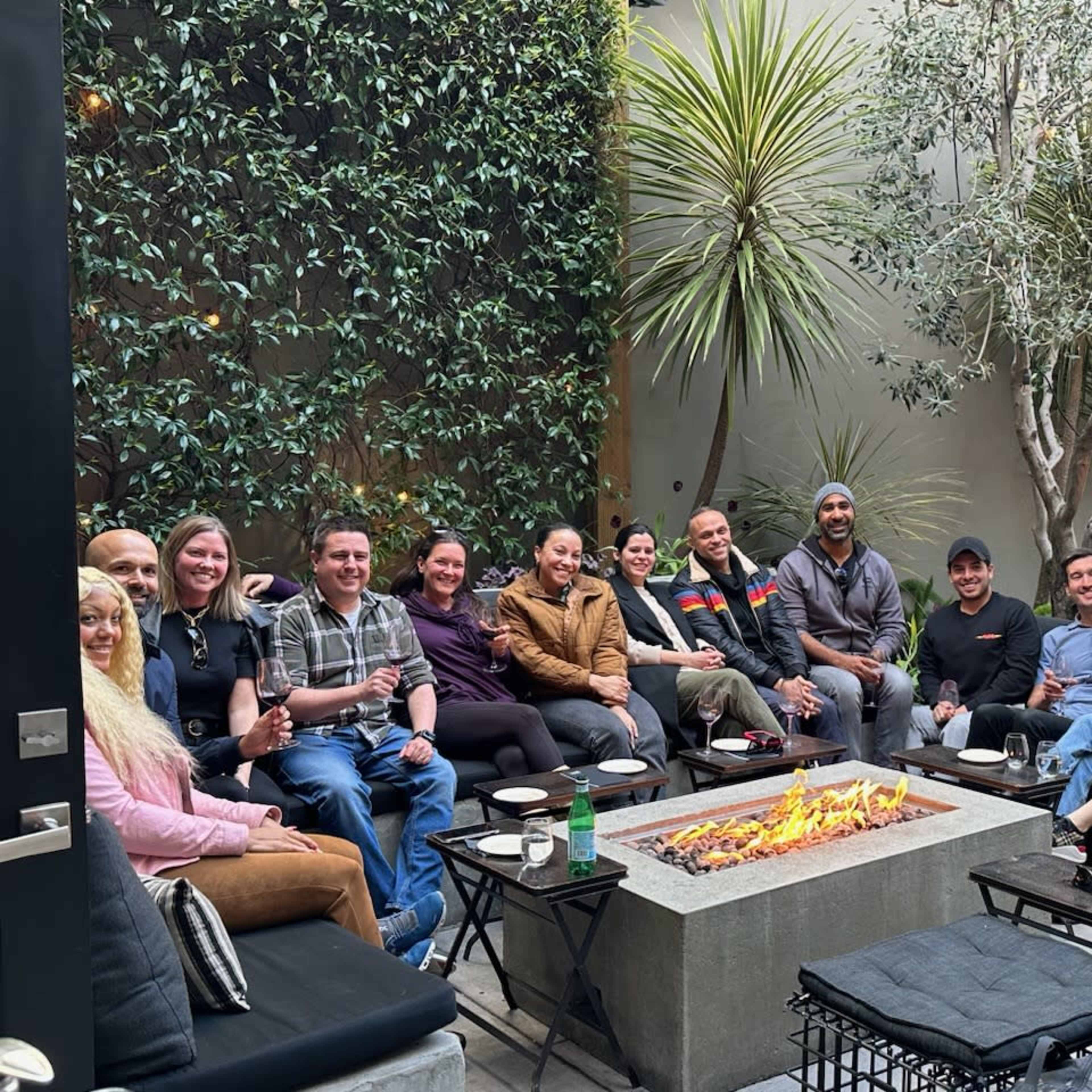 A group of people gathers around a fire pit in a lush outdoor seating area.