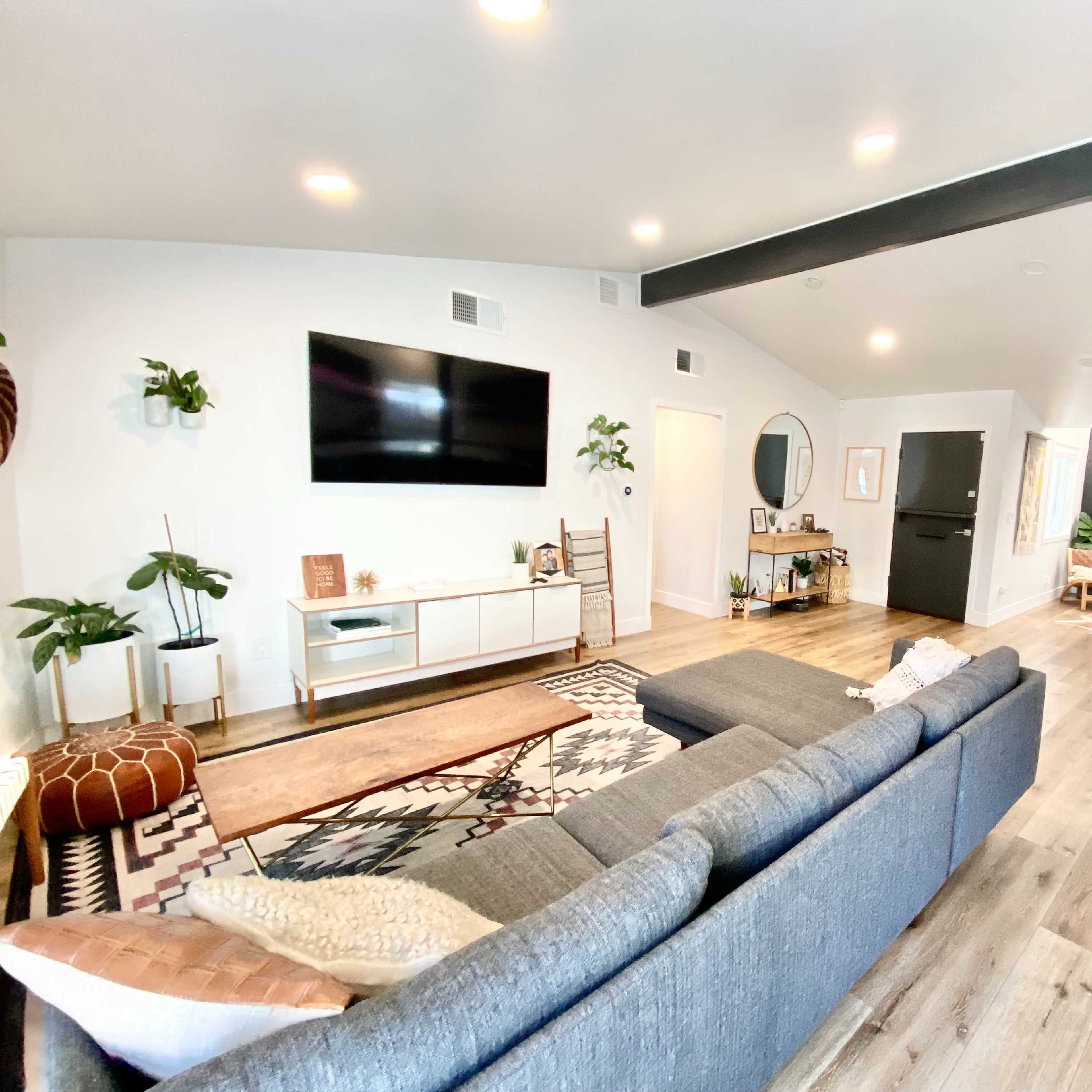 A modern living room with a sectional couch, a large television on the wall, and a dining area visible in the background.