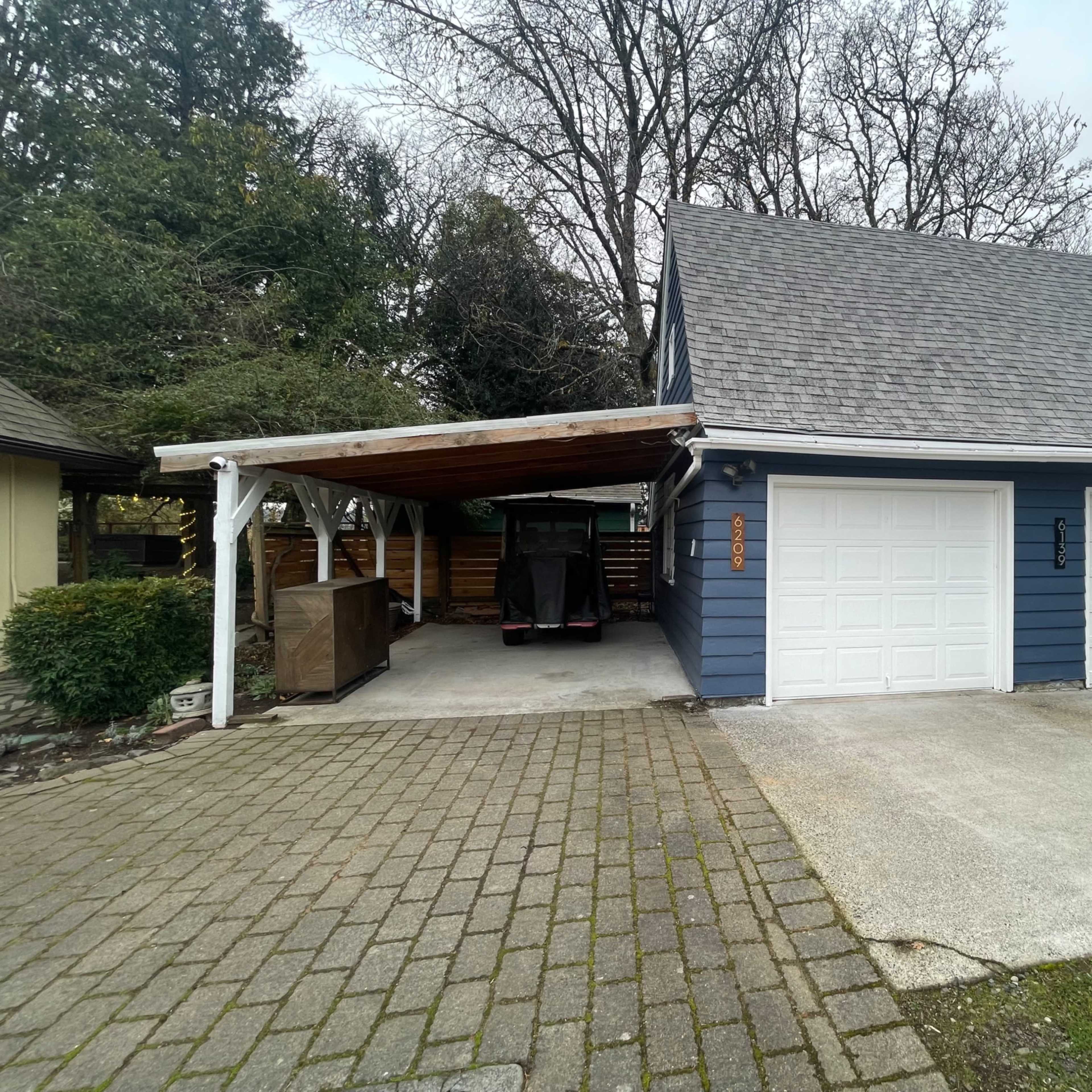 A blue house with a gabled roof features a two-car garage and a covered carport on a paved driveway.