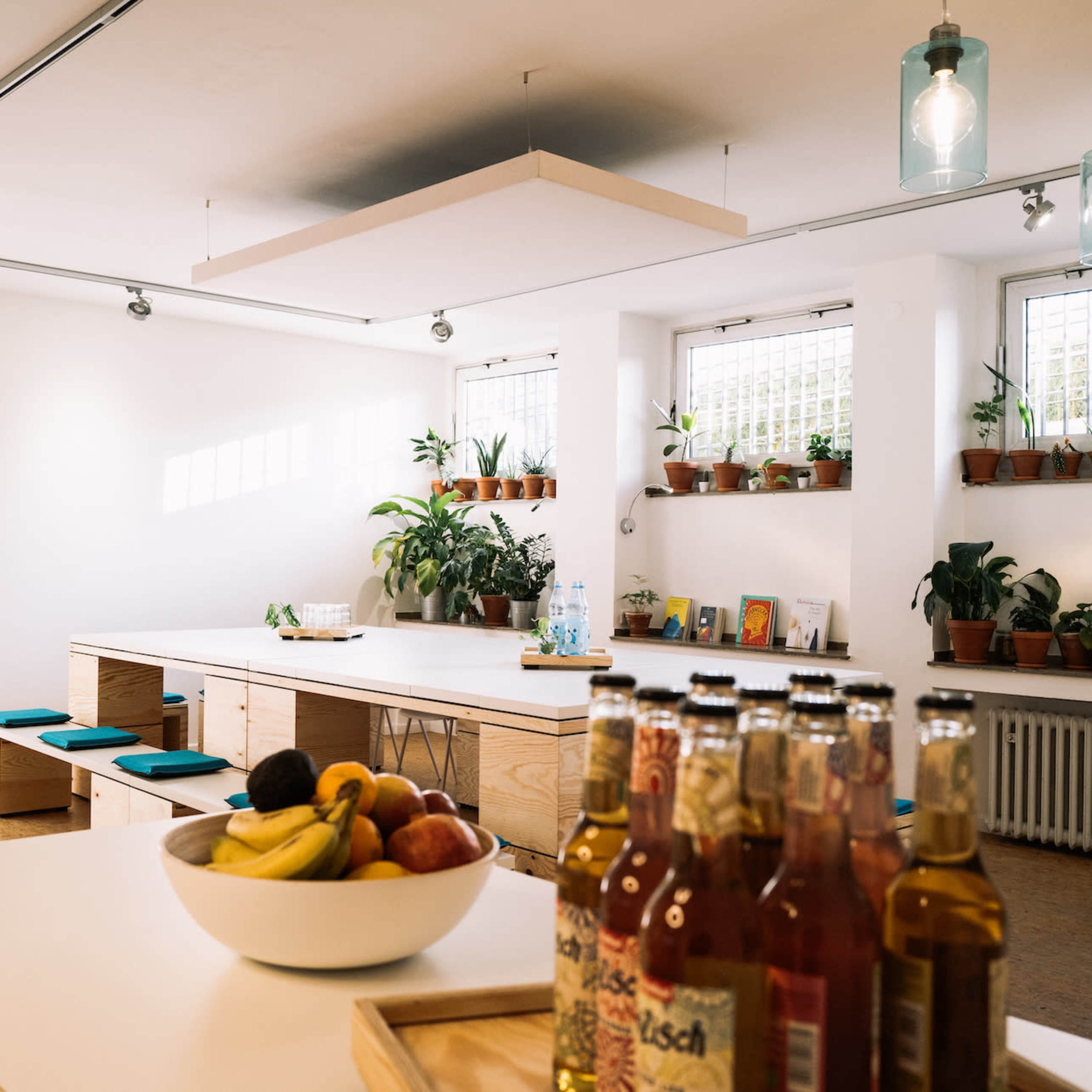 A modern kitchen and dining area features a long wooden table surrounded by potted plants and natural light streaming through the windows.