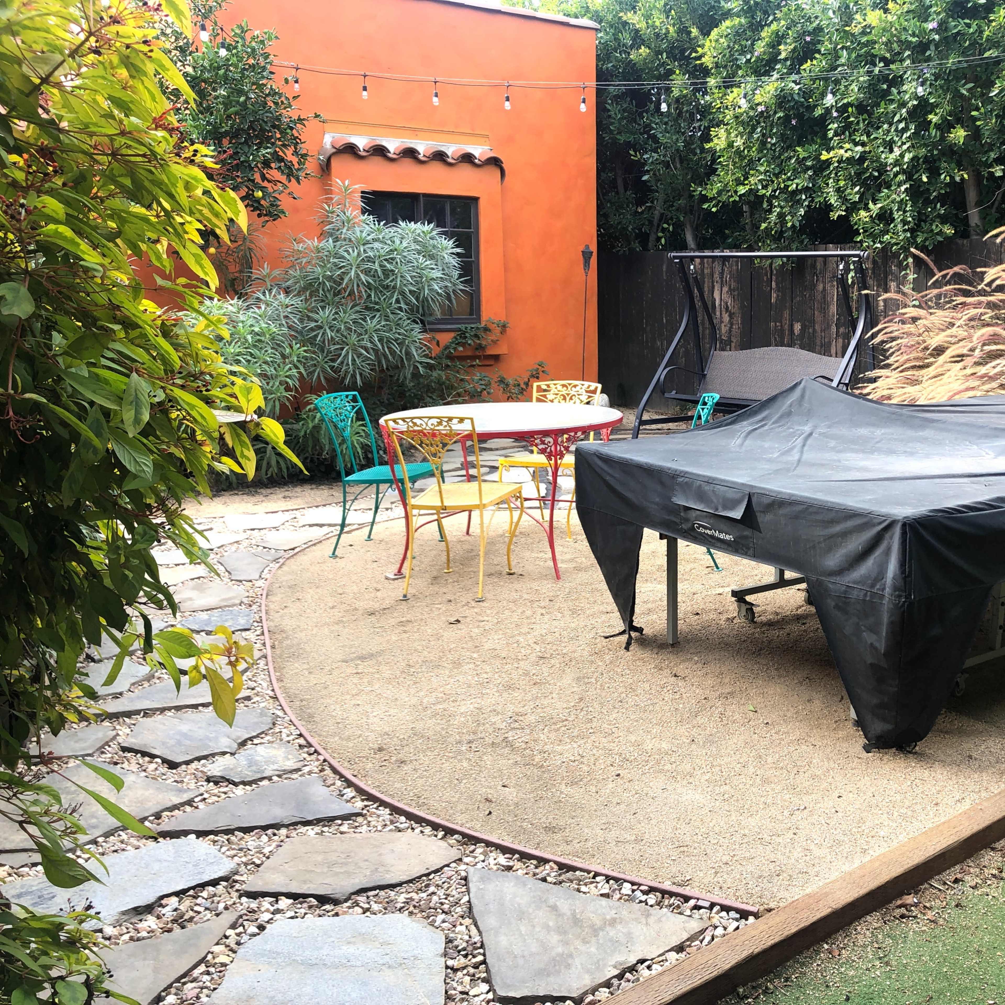 A backyard with a covered table and chairs, a gravel path, and surrounding greenery.