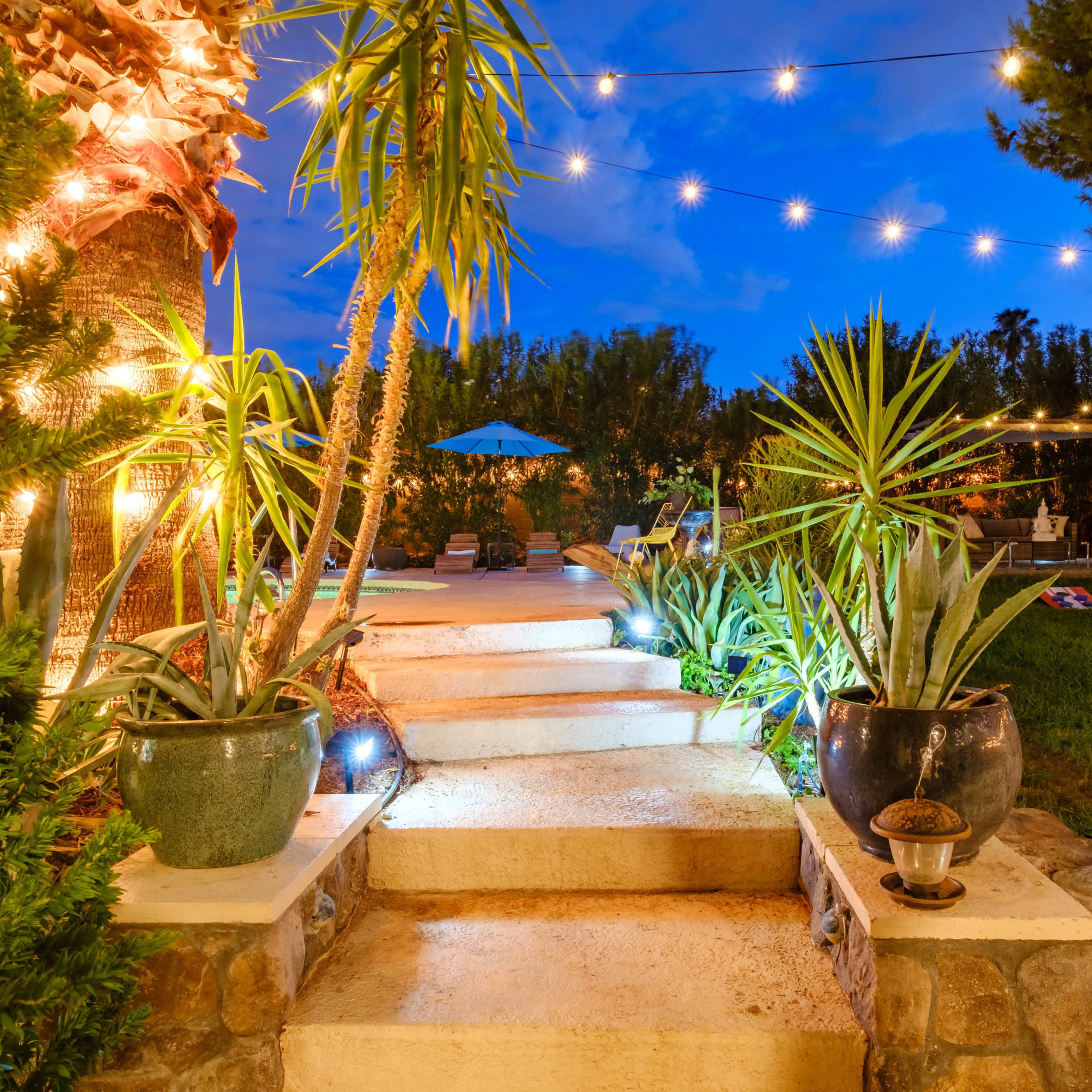 A set of well-lit stone steps leads through lush plants to a patio area illuminated by string lights at dusk.