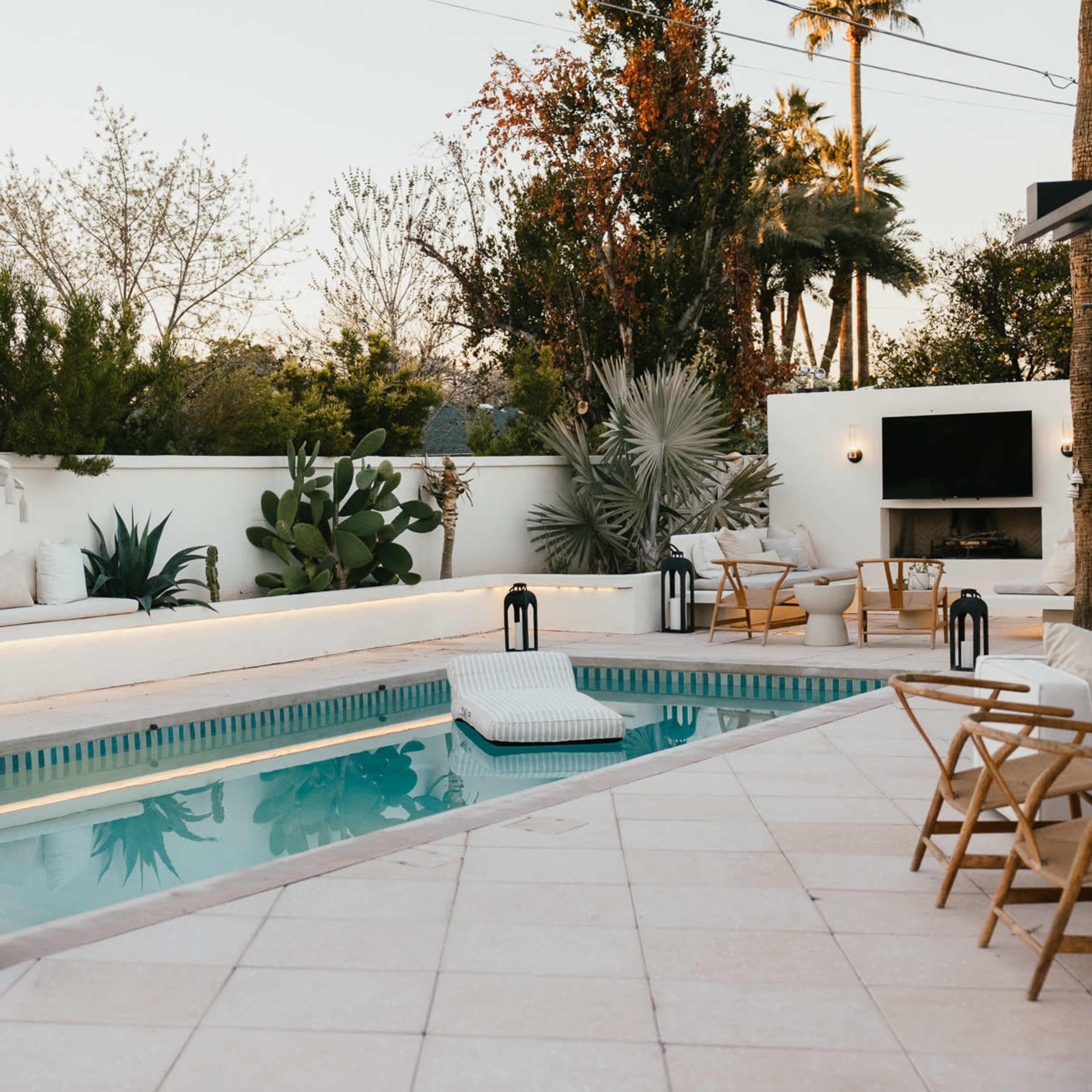The scene features a landscaped outdoor setting with a rectangular pool surrounded by lounge furniture, plants, and a large umbrella.