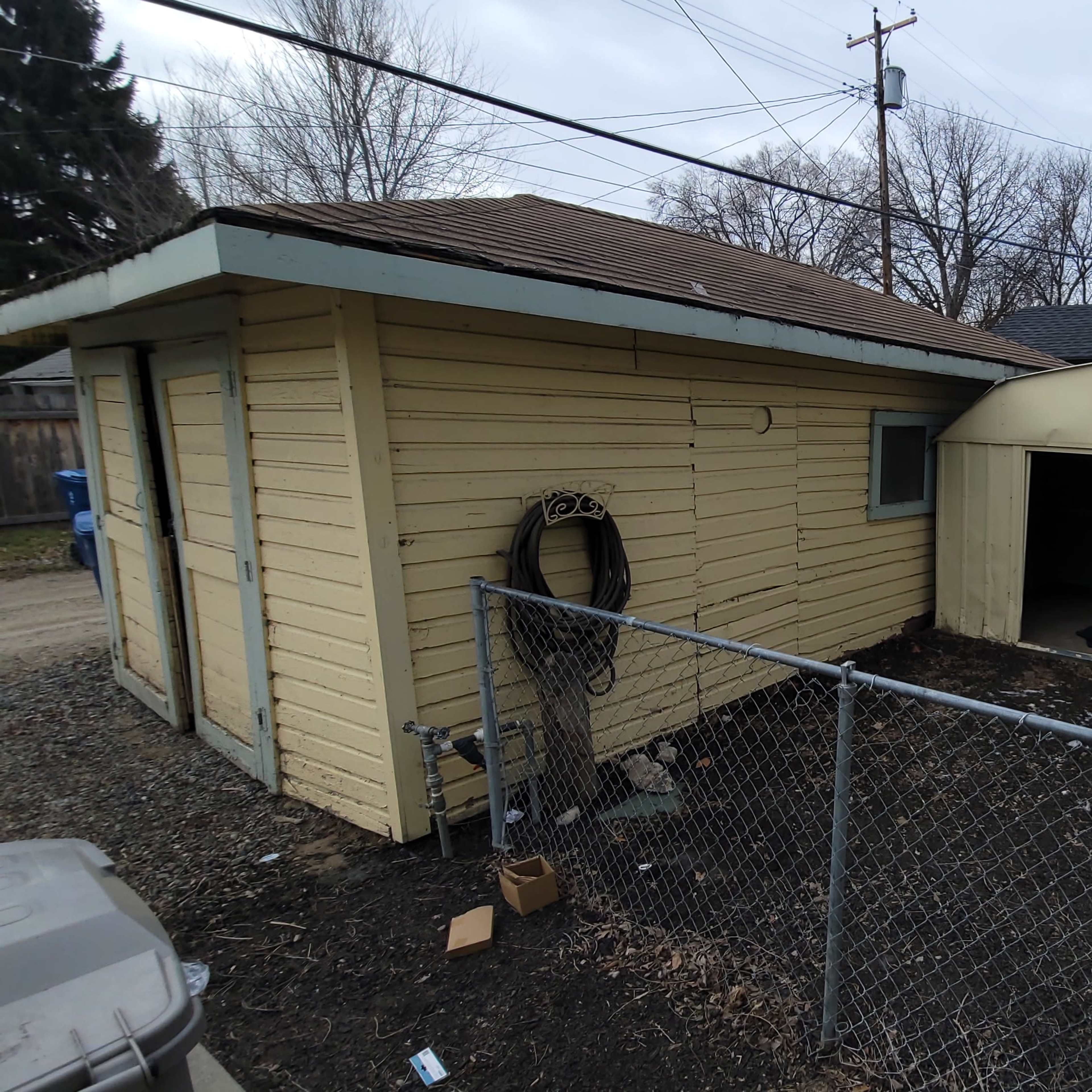 A yellow house with a brown roof sits next to a metal shed, enclosed by a chain-link fence in a yard with sparse grass and debris.