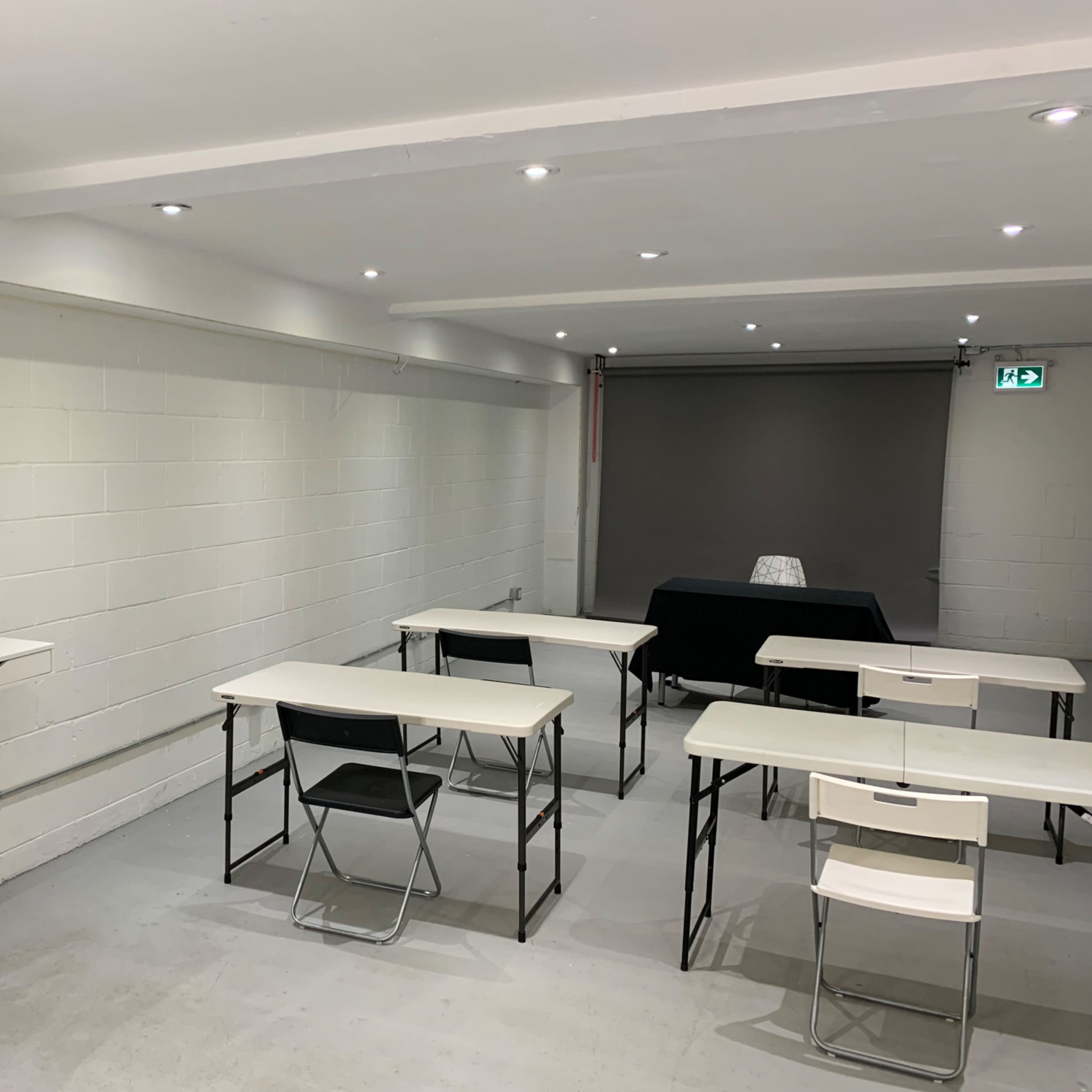 A minimalistic classroom setting with several tables and chairs arranged in a row, a large mirror on one wall, and a dark backdrop along the opposite side.