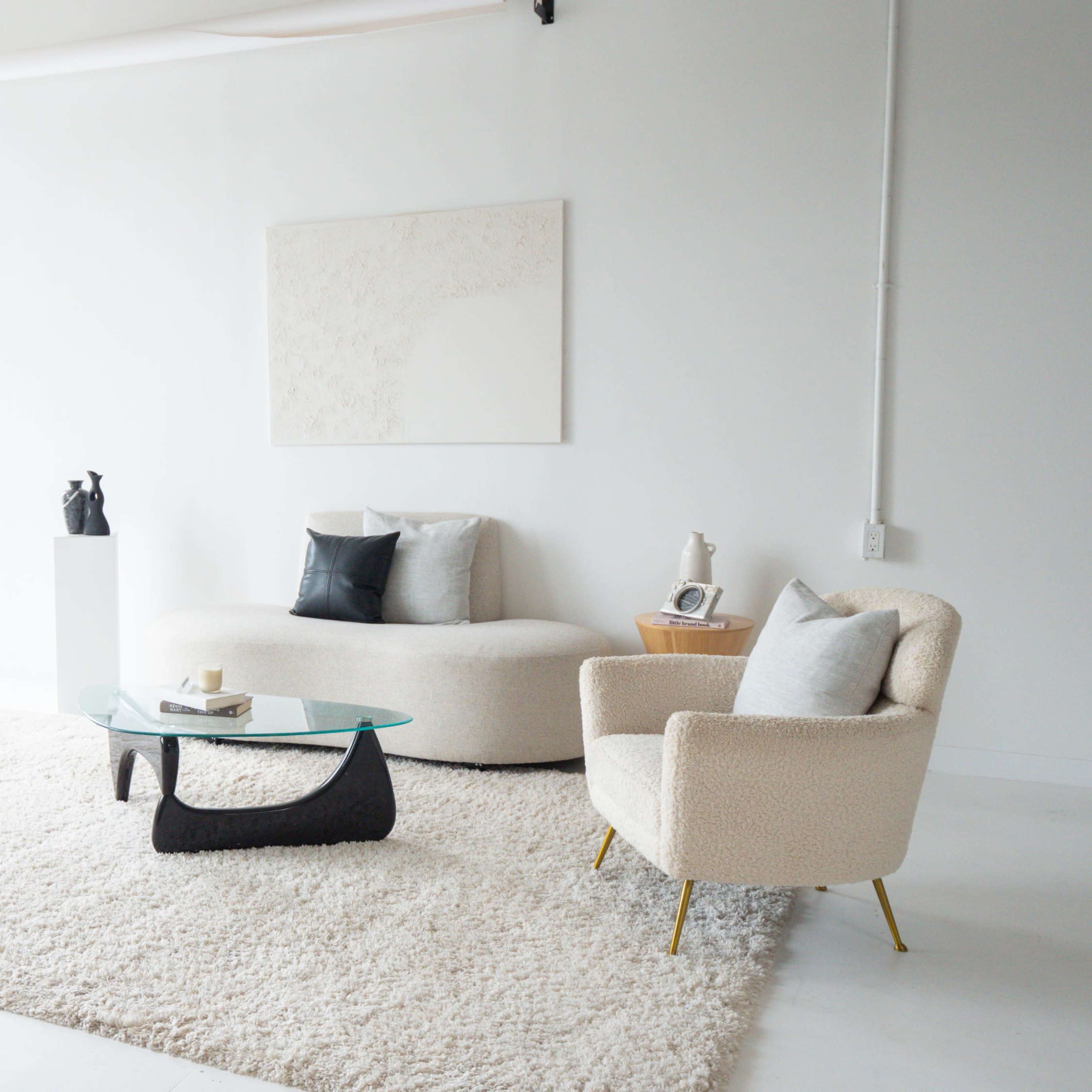 A minimalist living room features a light-colored sofa, an armchair, a glass coffee table, and a large area rug, all arranged around a bright, spacious window.