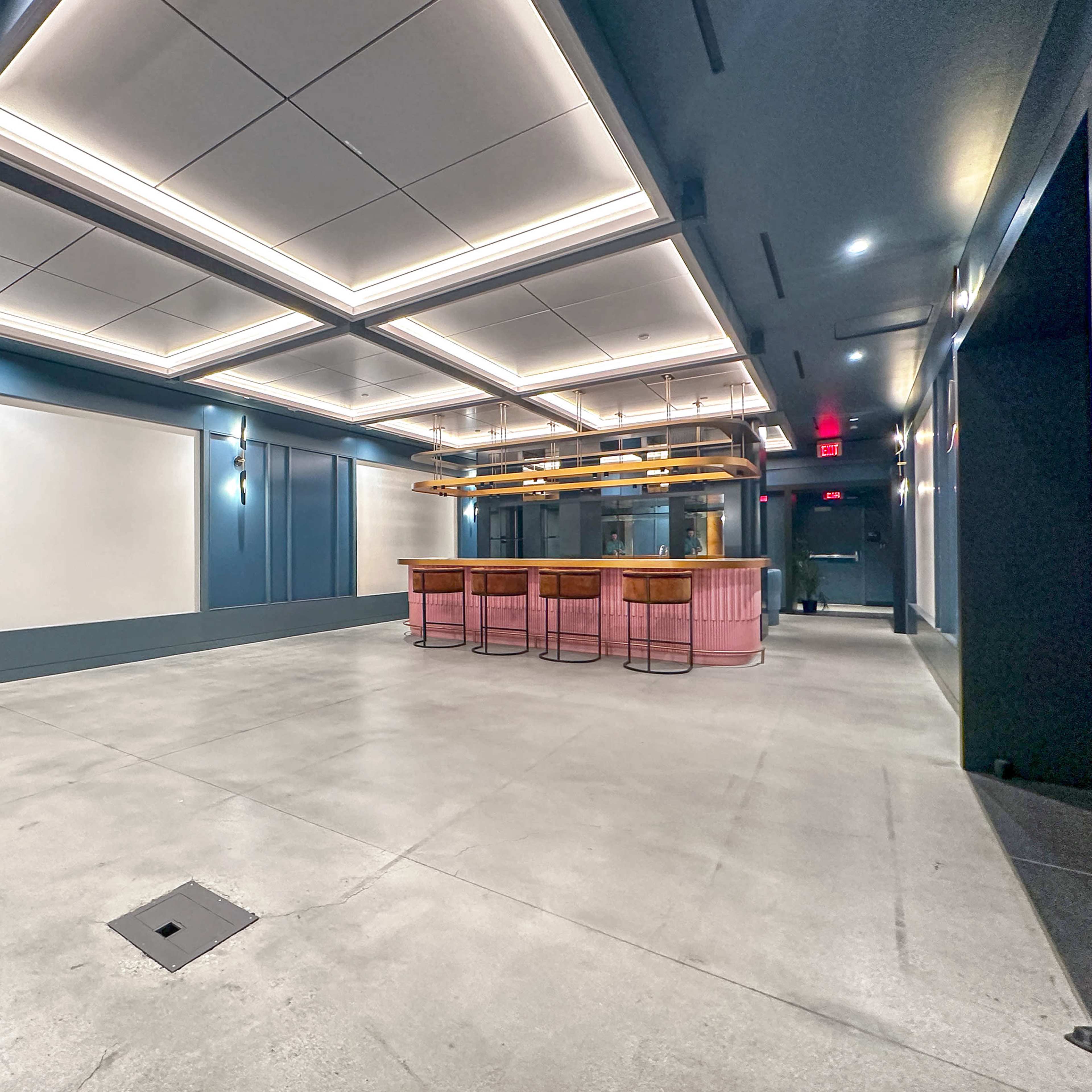 An empty modern space featuring a bar with wooden stools and overhead lighting against a backdrop of blue walls and concrete flooring.