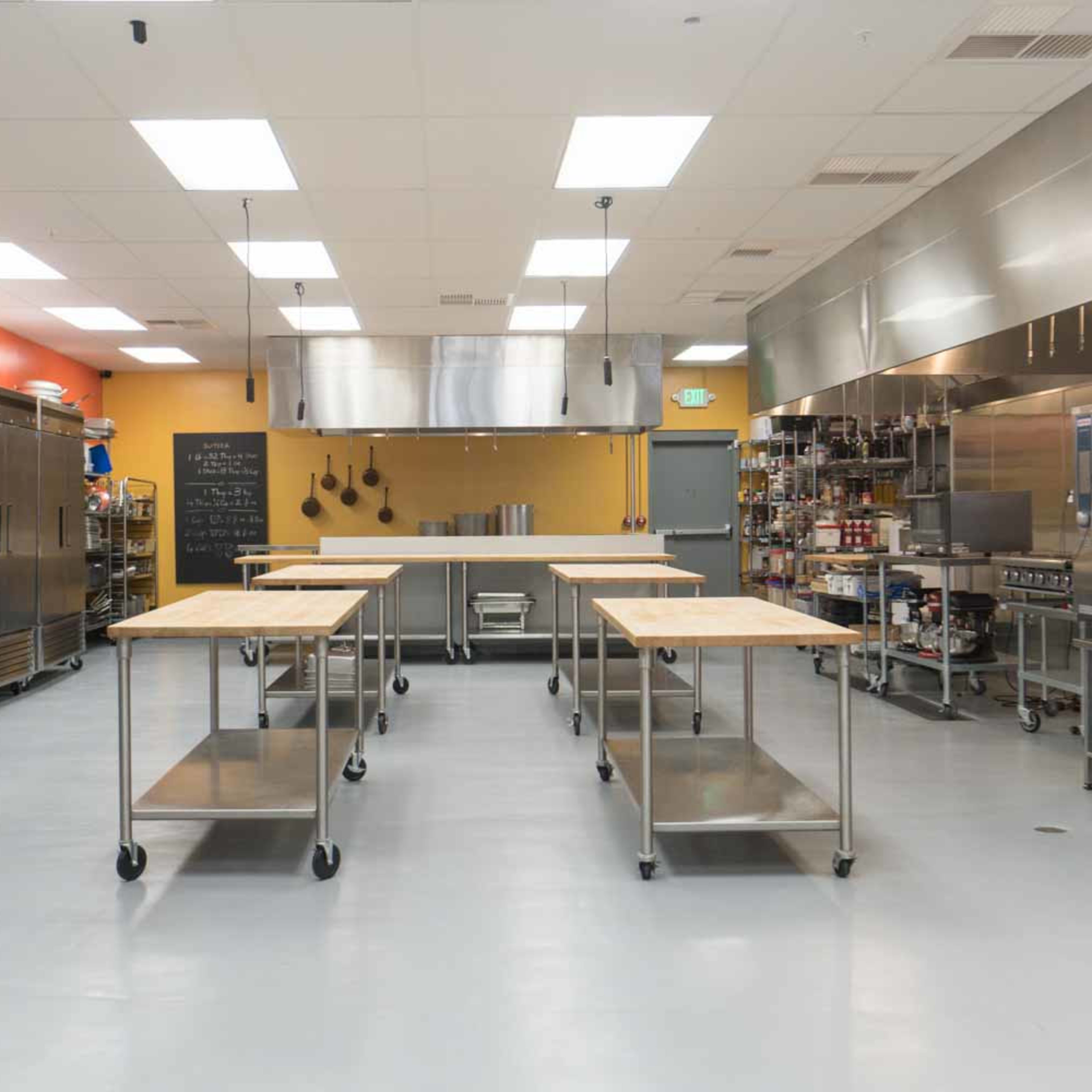A modern kitchen space with stainless steel appliances, several wooden prep tables, and well-organized storage areas on the walls.