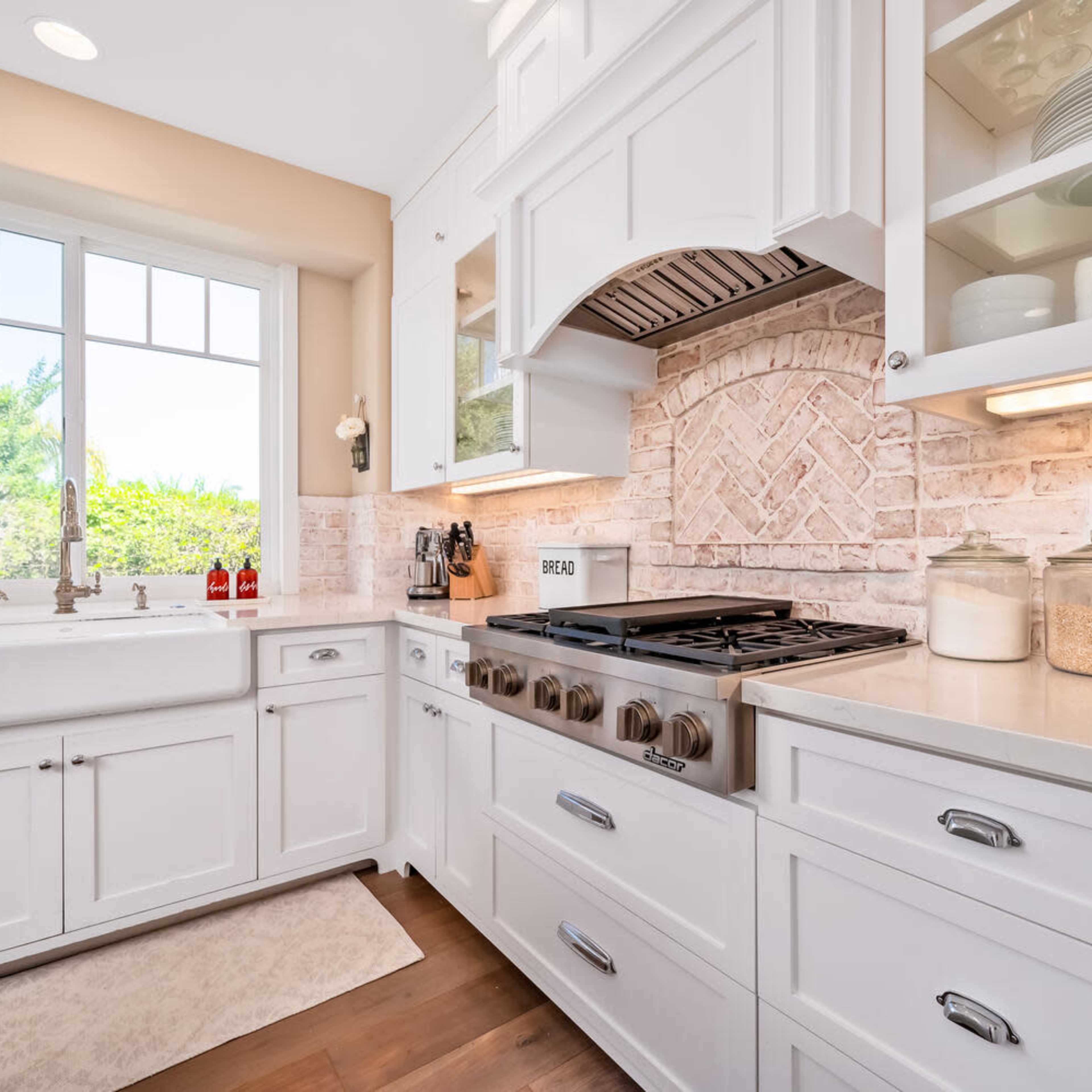 A well-organized kitchen with white cabinetry, a farmhouse sink, and a gas range under a decorative hood, along with jars of ingredients displayed on the countertop.