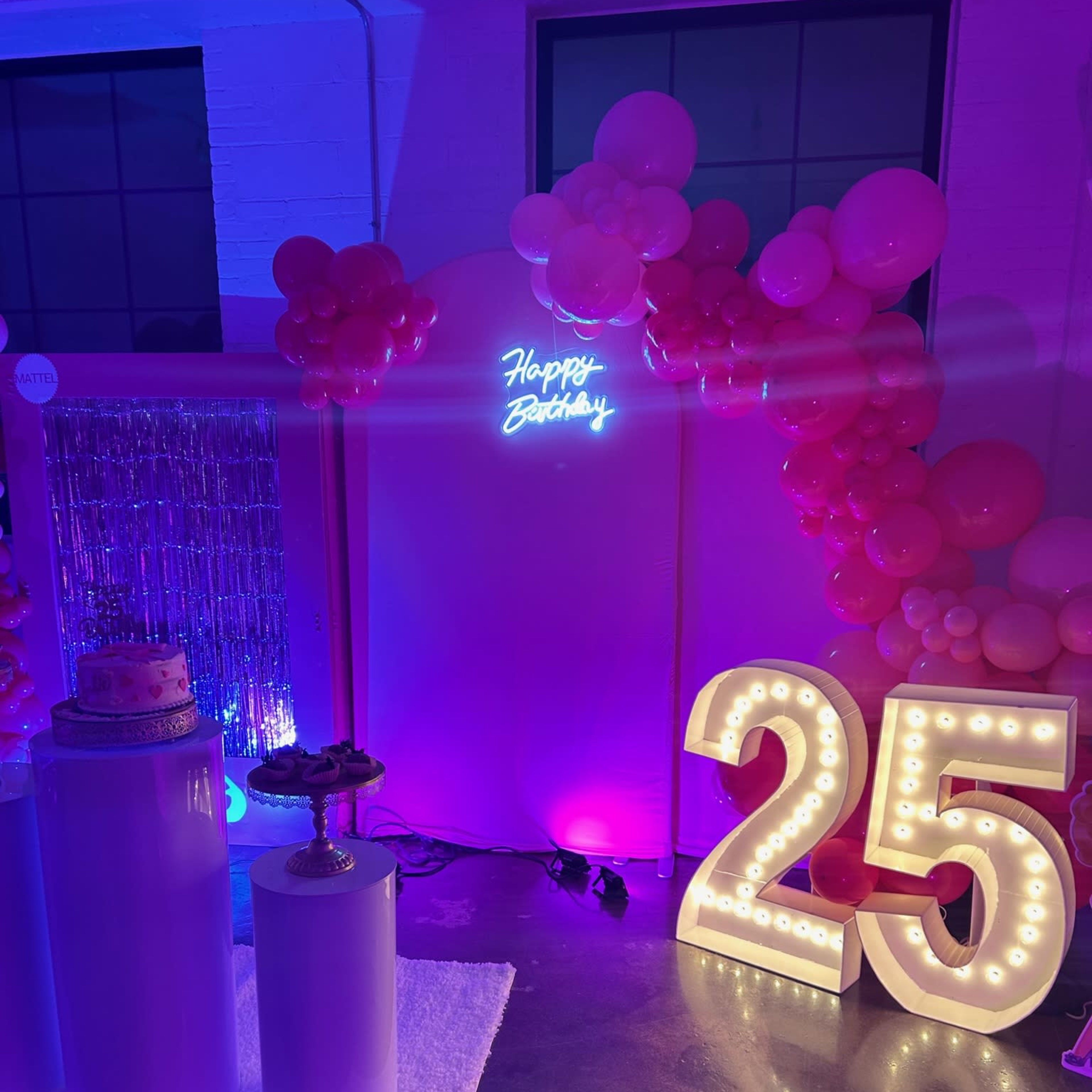 A festive birthday party setup featuring a large illuminated "25" sign, a neon "Happy Birthday" sign, and a decorated cake on a pedestal surrounded by pink balloons.