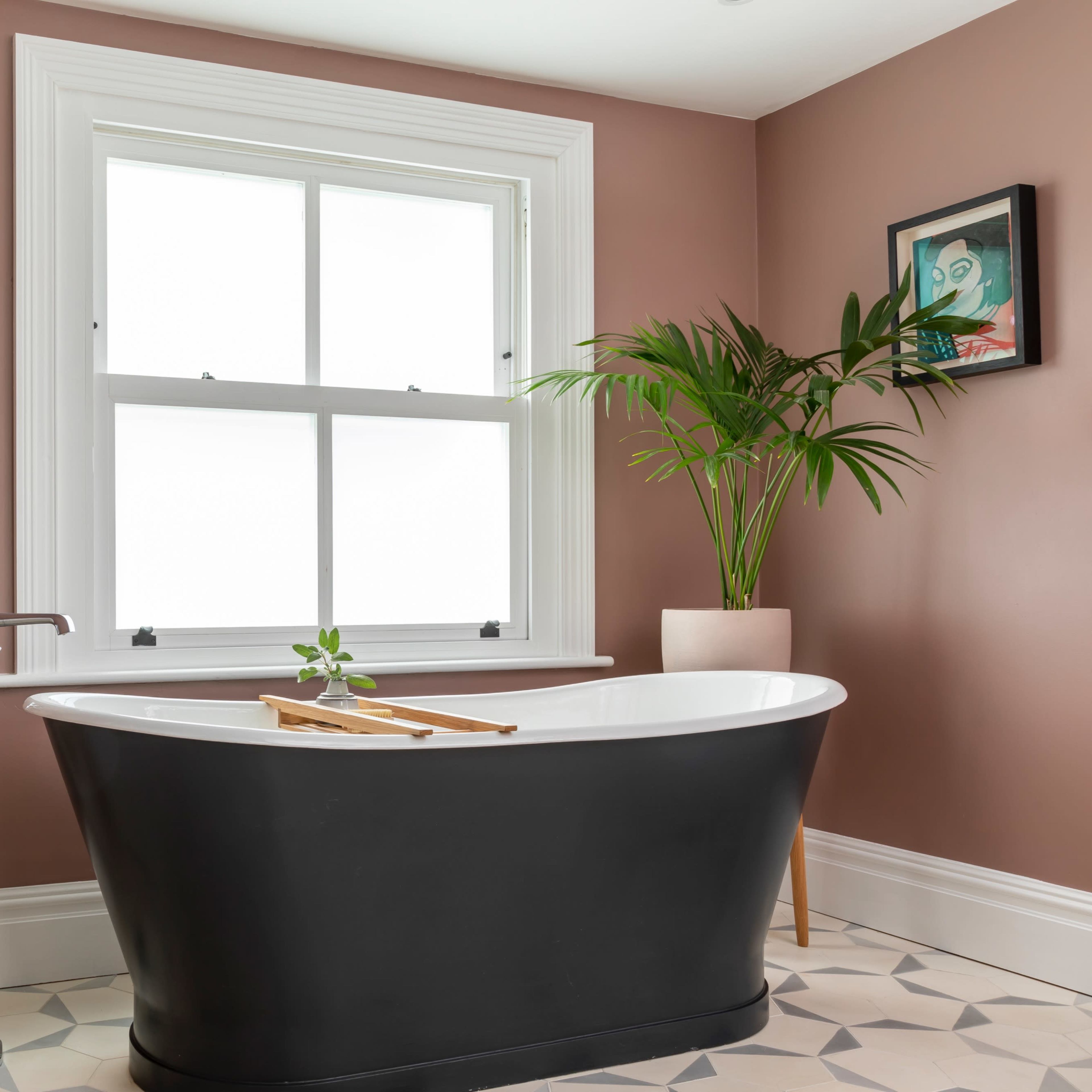 A black freestanding bathtub is positioned beside a large window in a bathroom featuring a beige wall and a plant in a pot.