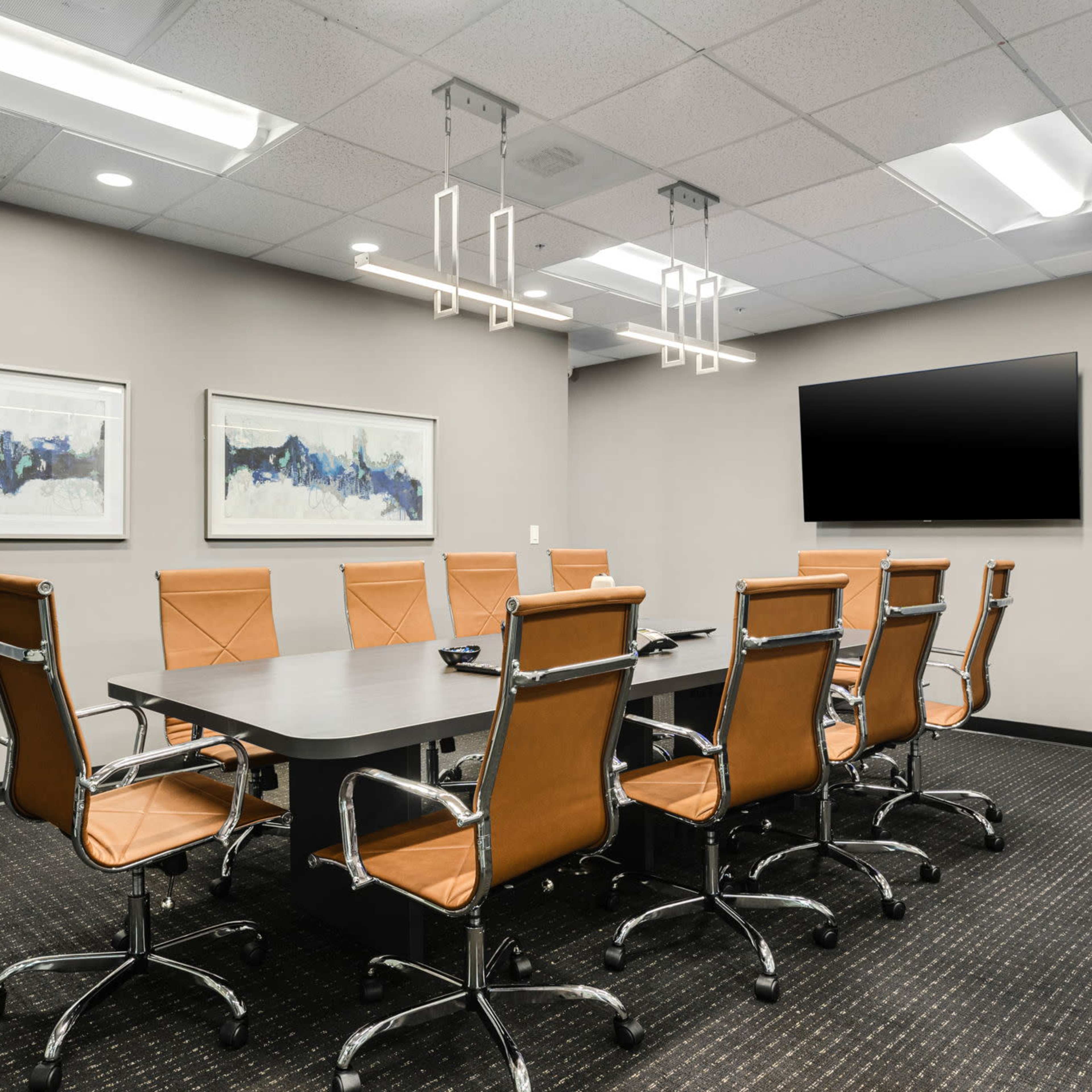 A conference room features a long table surrounded by eight brown leather chairs, with two abstract paintings on the wall and a wall-mounted television.