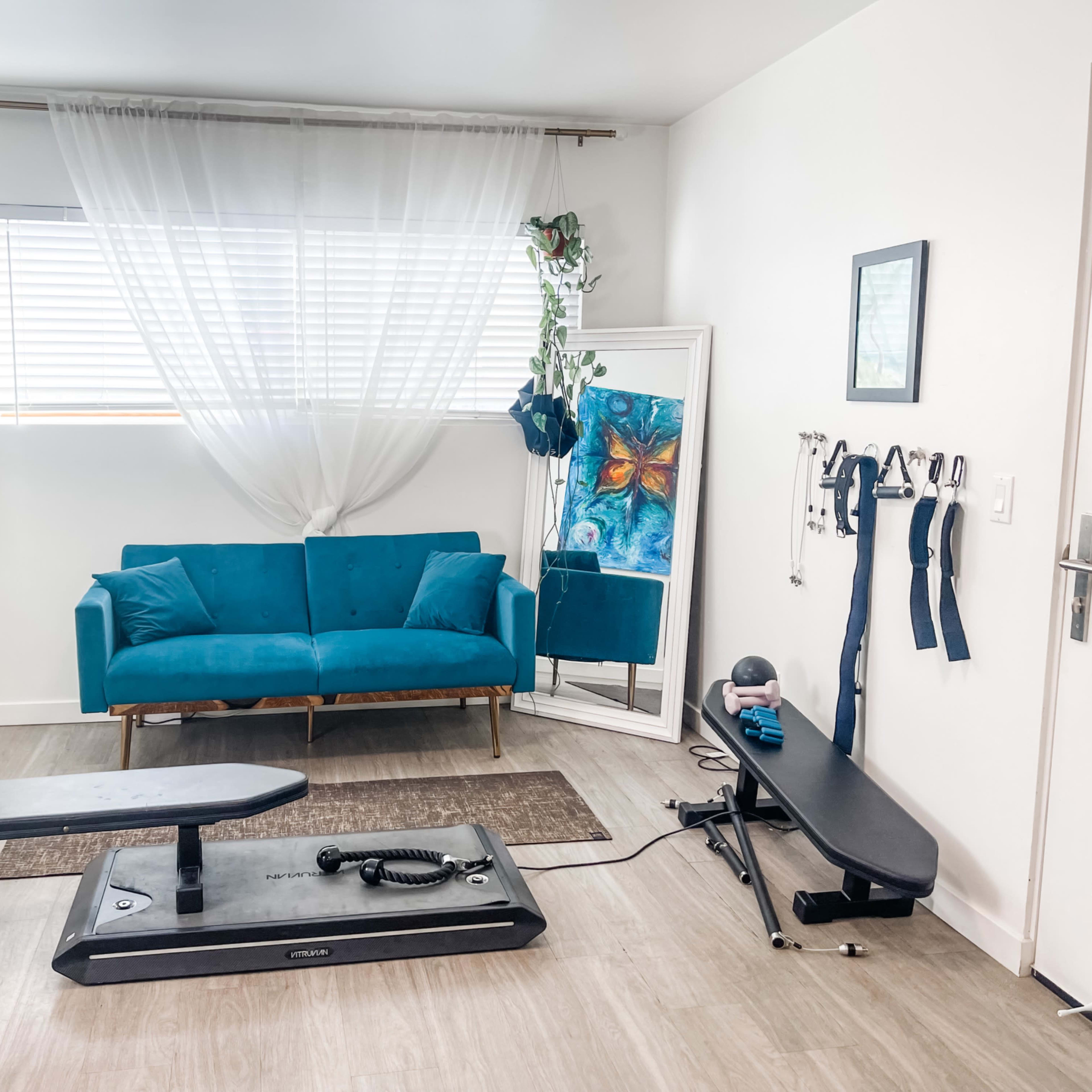 A fitness area features a blue couch, a mirror, and exercise equipment including a vibration plate and resistance bands against a white wall with a window.