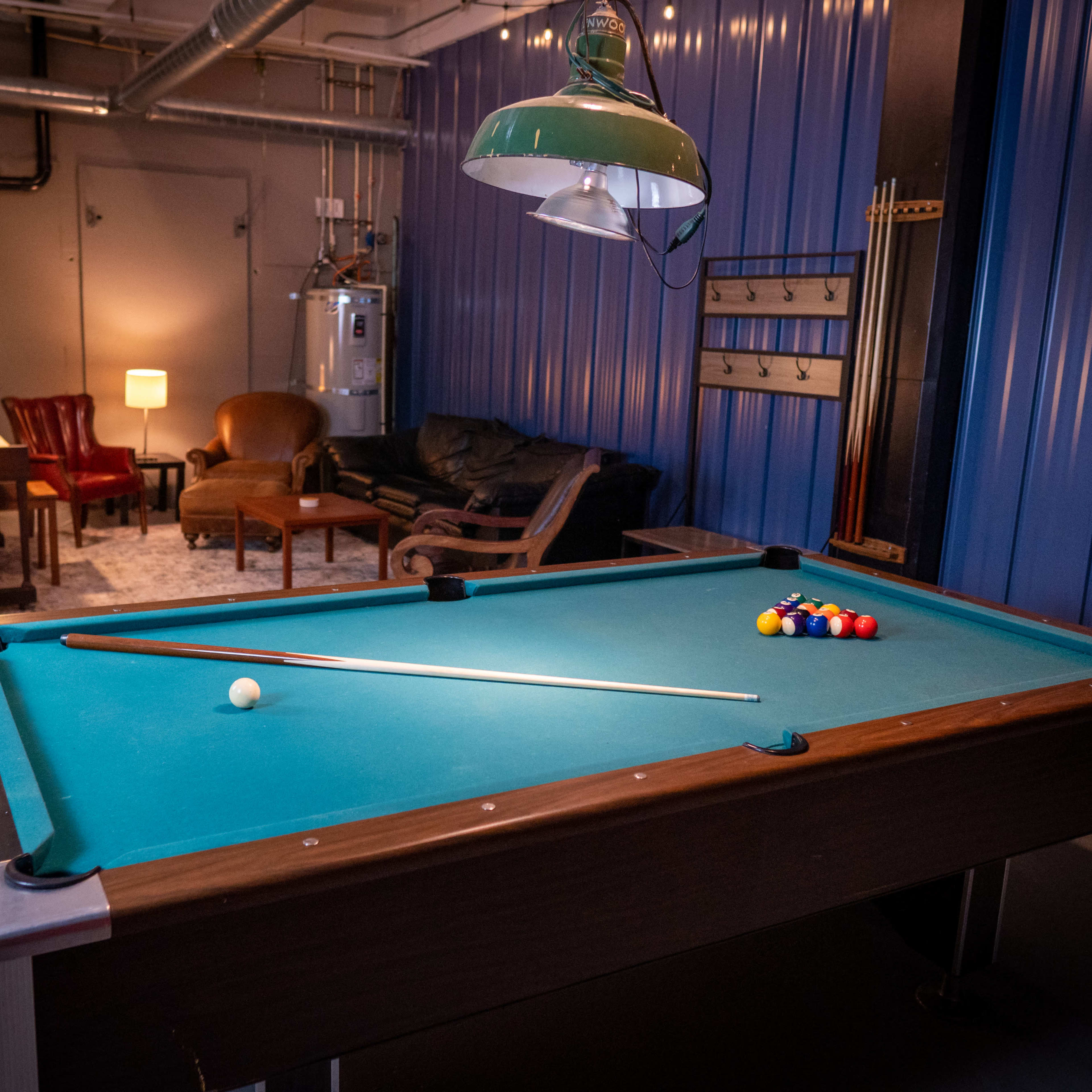 A well-lit room features a billiard table with a cue stick and balls arranged in a triangle, alongside a seating area with a couch and armchair.