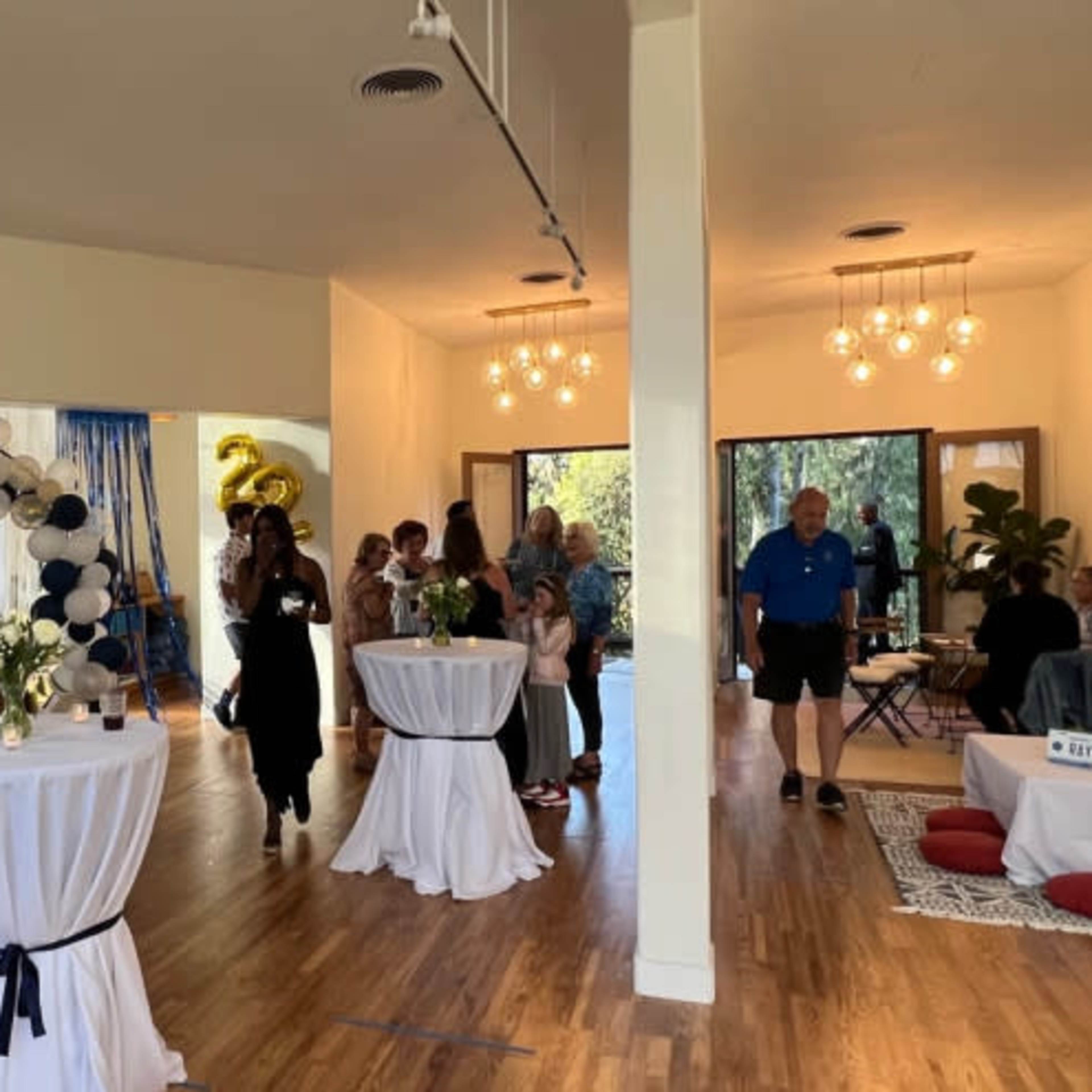 A group of people socializes at a spacious indoor event, featuring tables with refreshments and a decorative balloon arch in the background.