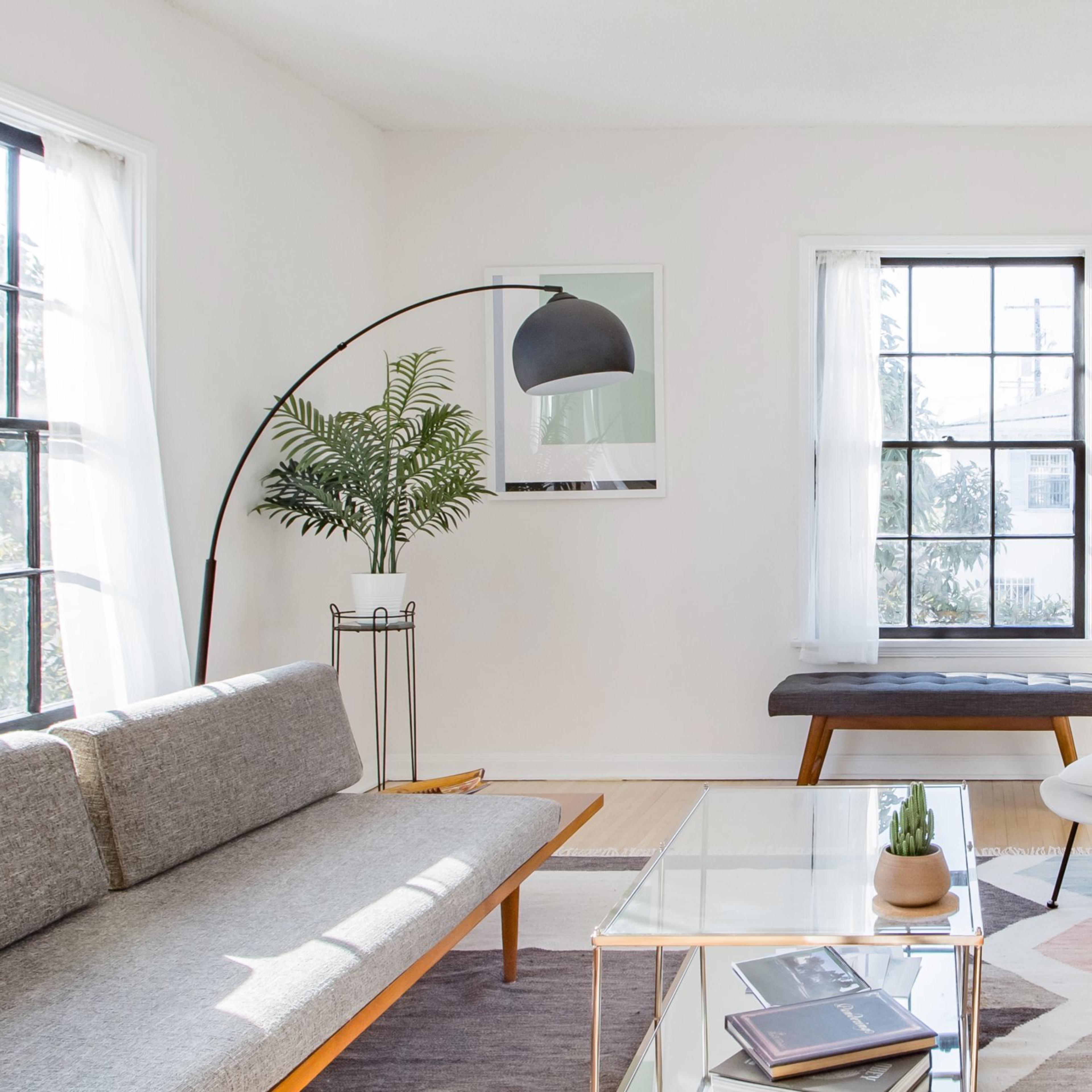 A modern living room features a gray sofa, a glass coffee table, a light chair, and large windows with a view of greenery outside.