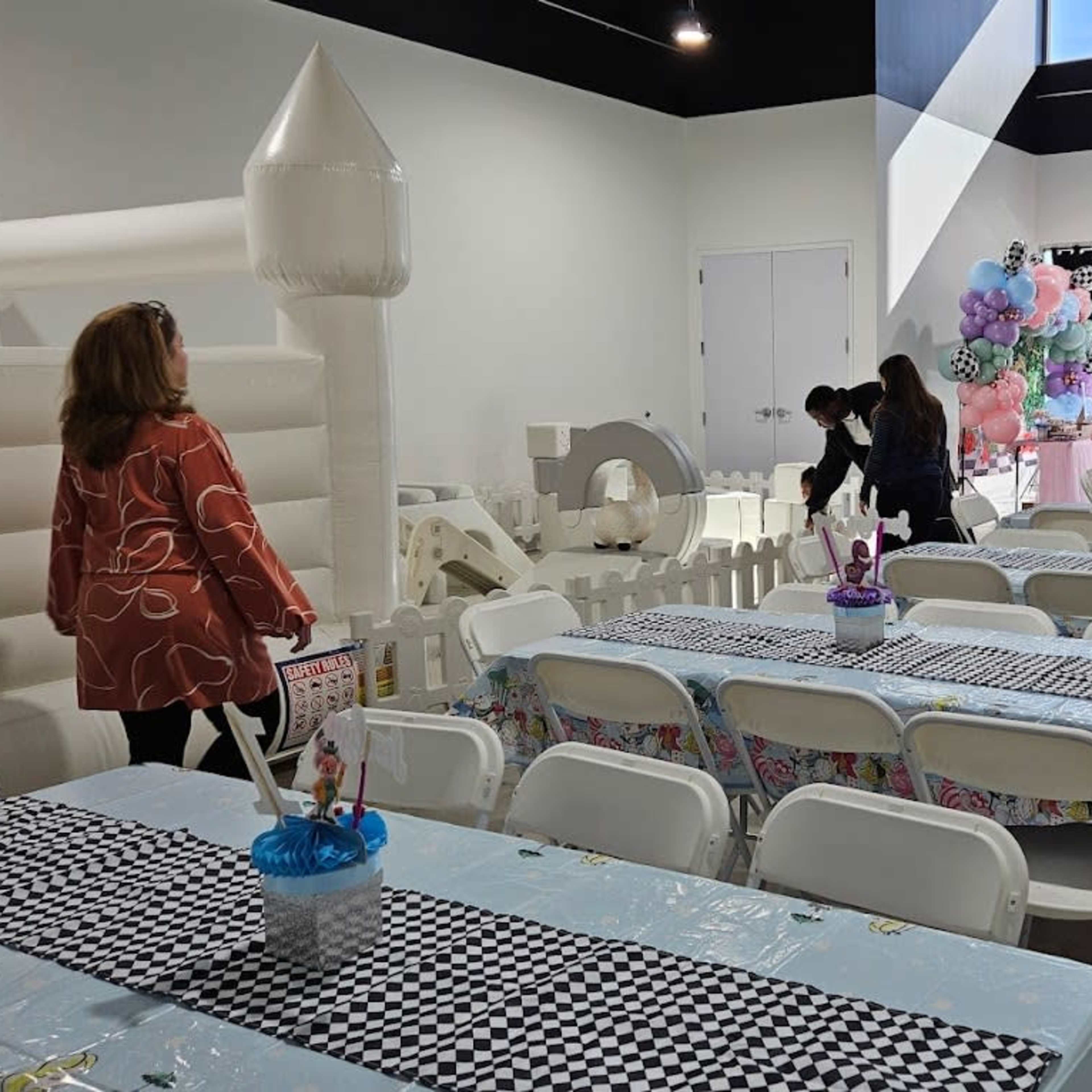 A spacious indoor venue set up for a party, featuring rows of tables with colorful tablecloths, chairs, and a large inflatable structure in one corner.