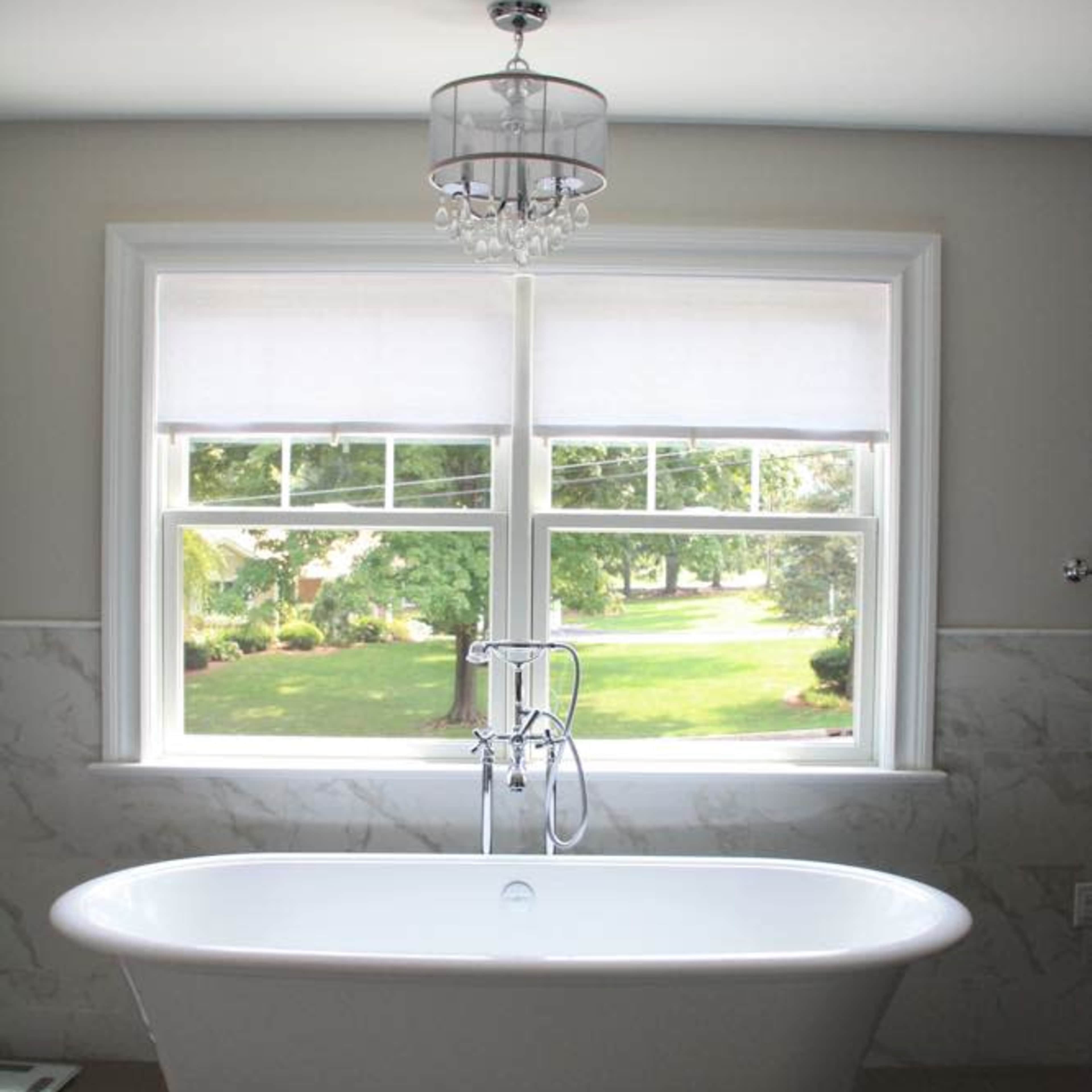 A freestanding white bathtub is positioned under a window that offers a view of a green landscape, illuminated by natural light.