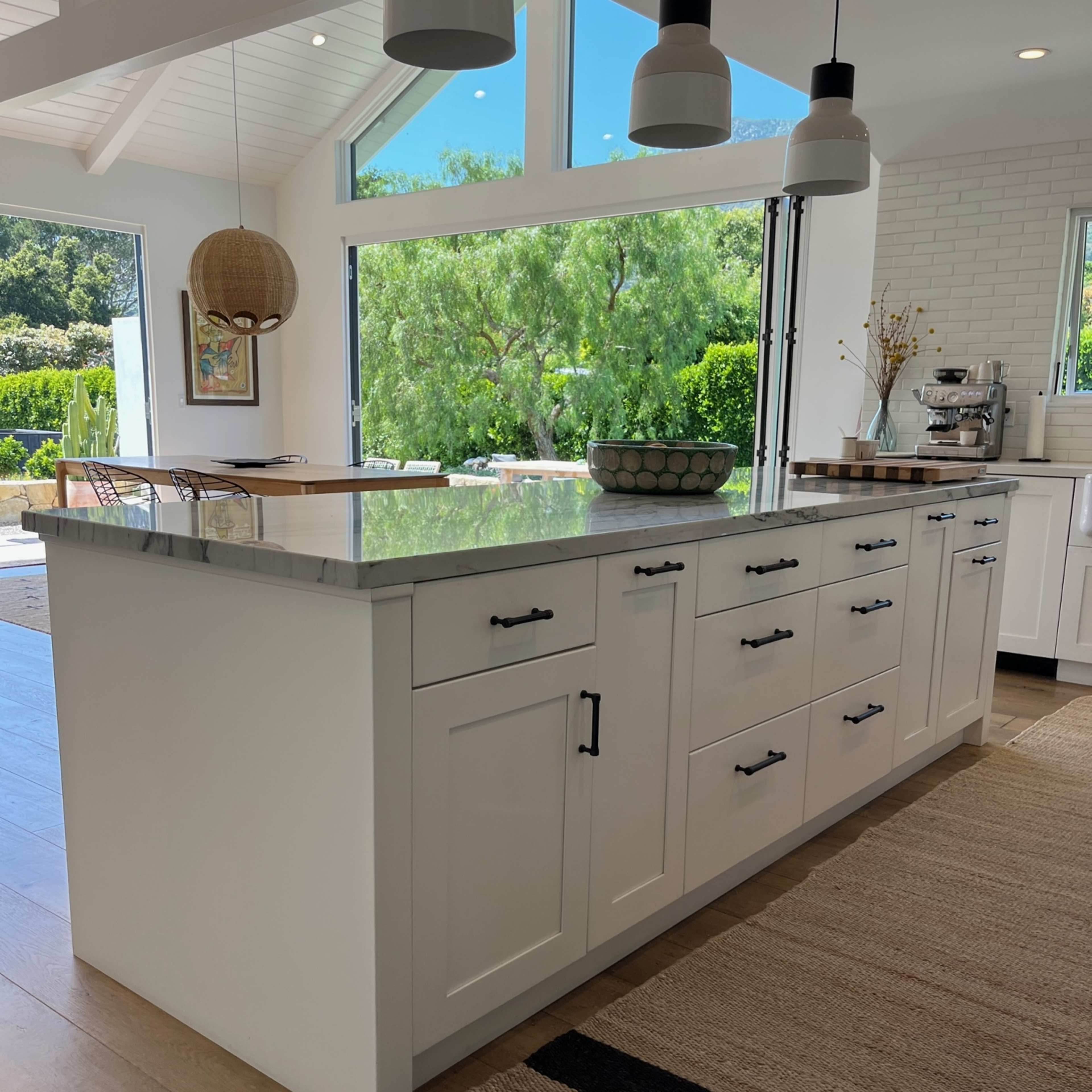A modern kitchen features a large island with white cabinets and a countertop, surrounded by natural light from large windows.