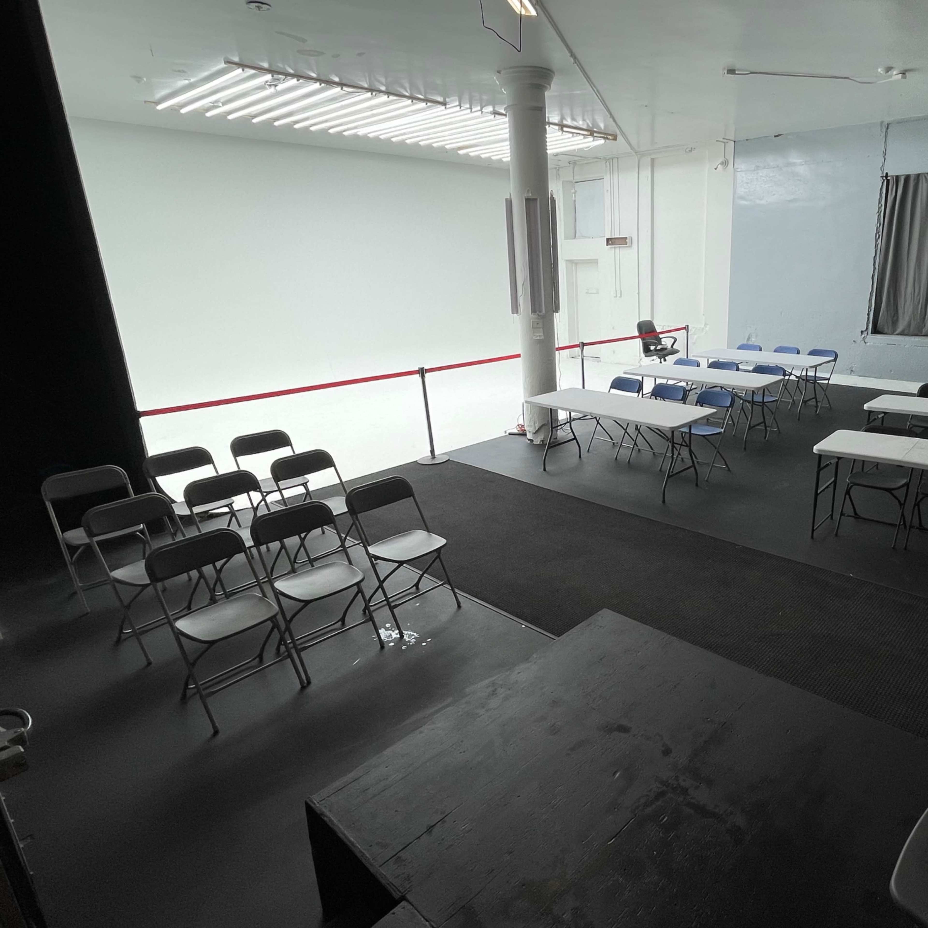 An empty room with multiple tables and chairs arranged in a setup for a meeting or event, with a white wall and a gray window in the background.