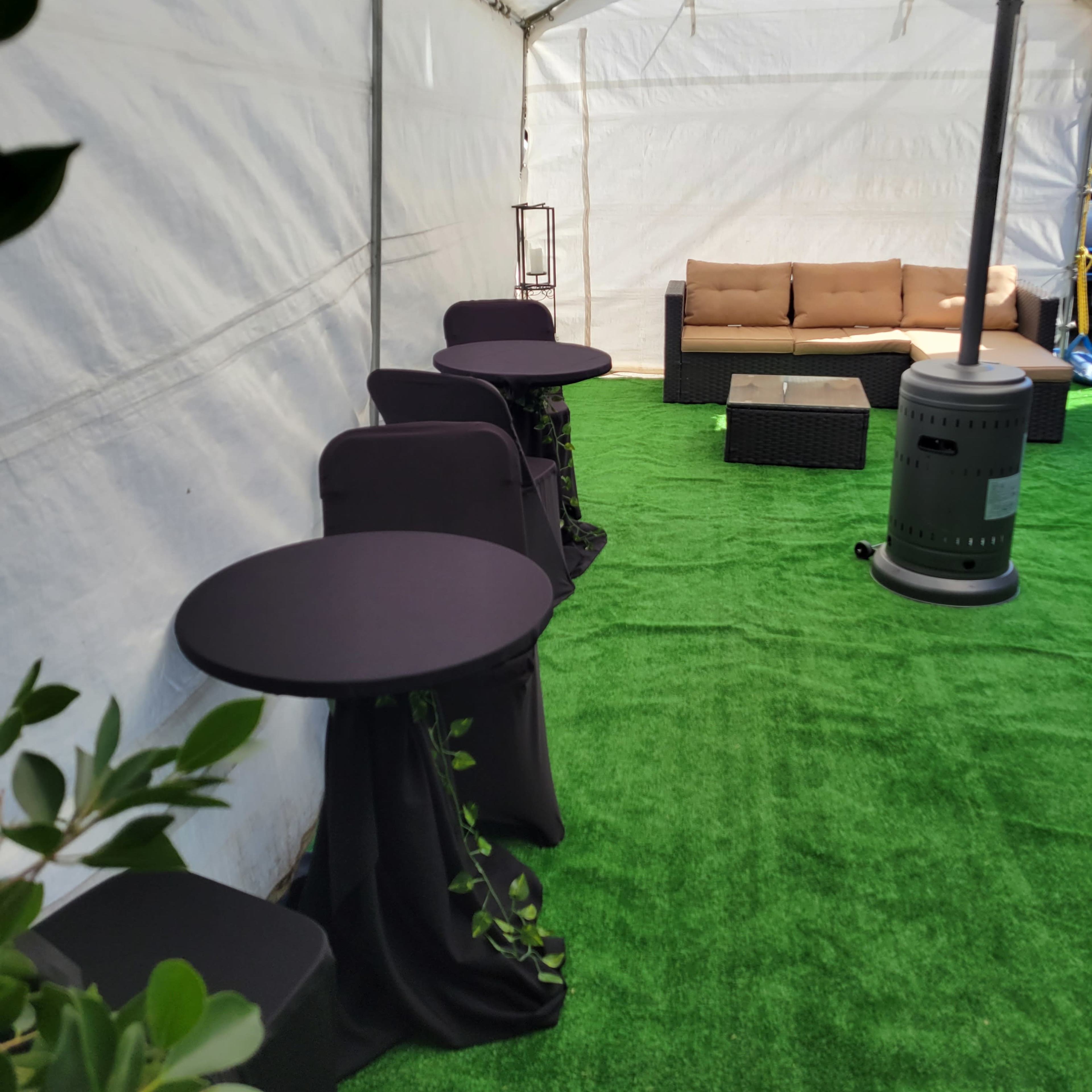 An indoor tent setup with black table covers, green artificial grass flooring, and a brown seating area along one side.