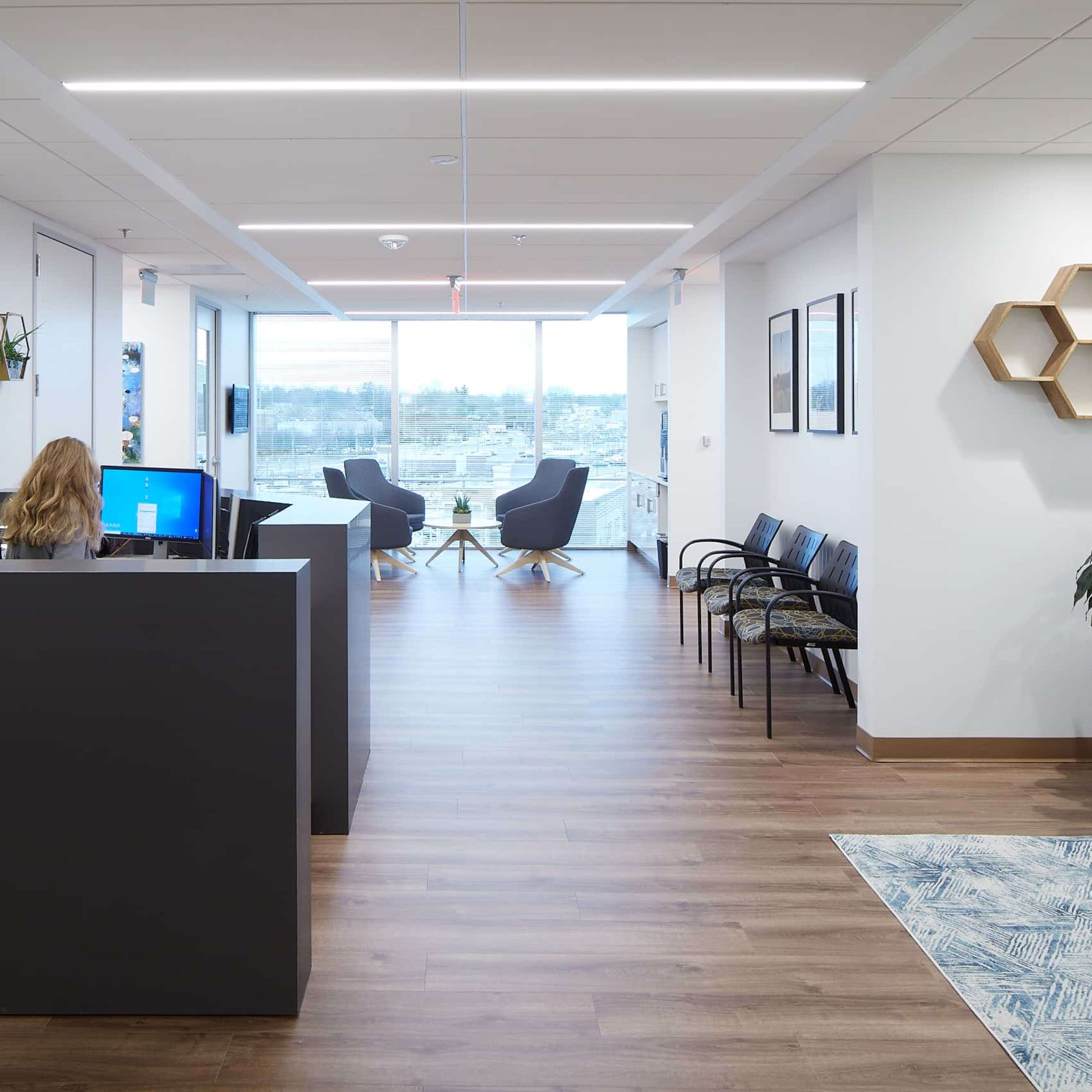 An office reception area features a desk with a computer, chairs along the wall, and large windows overlooking a cityscape.