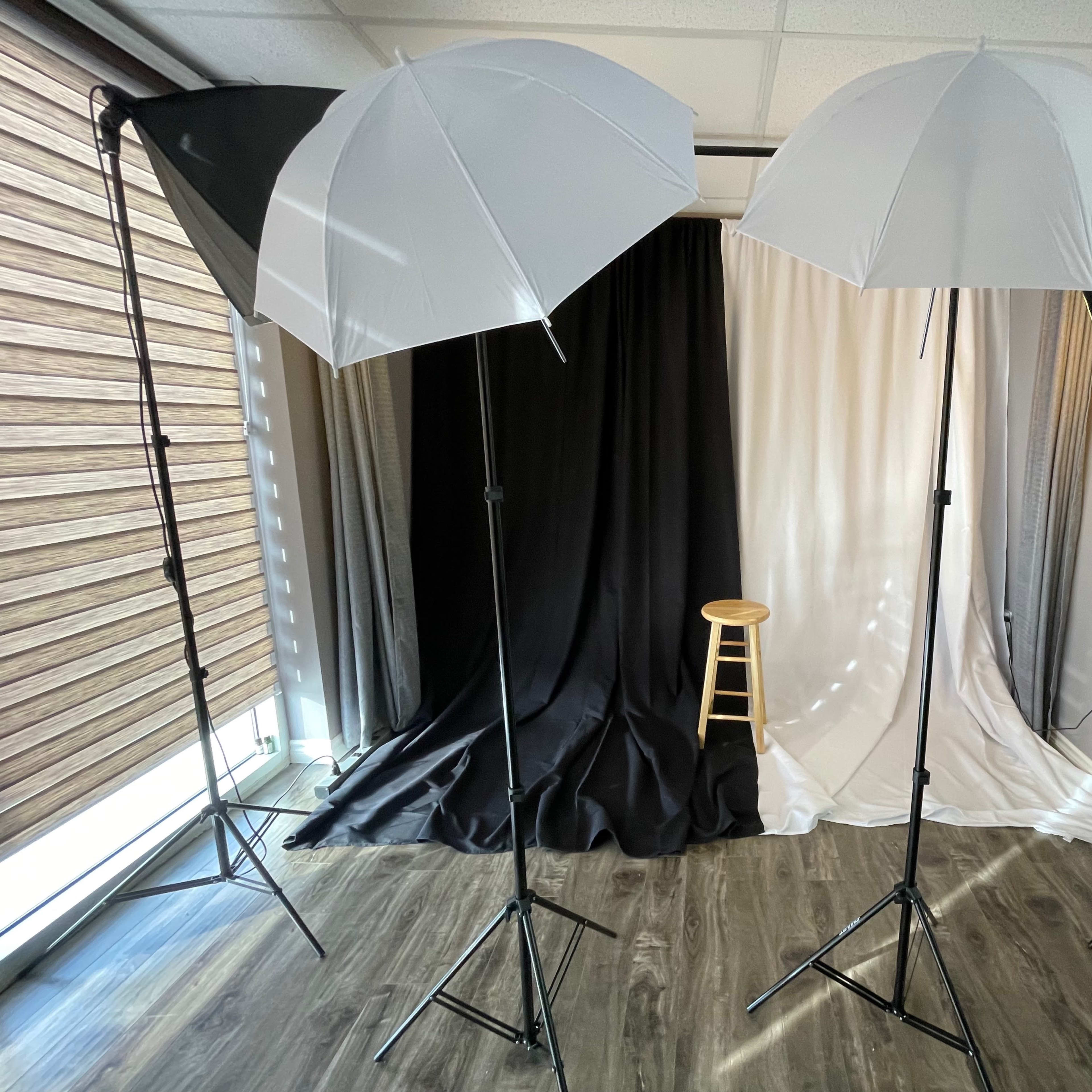 A well-lit photography setup with two umbrella lights on stands facing a three-panel backdrop of black, white, and gray, with a wooden stool positioned in front.