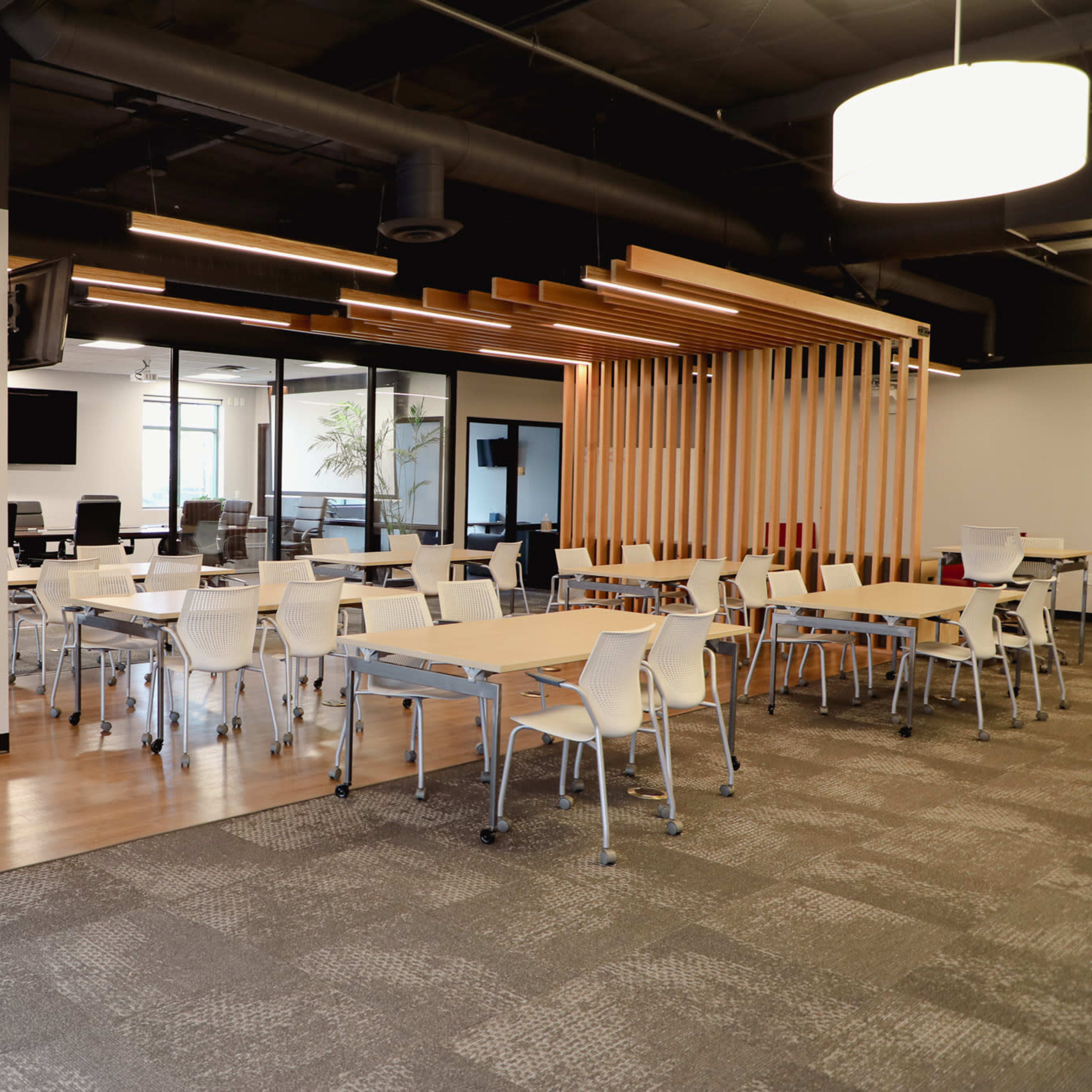 A modern office space with several tables and chairs arranged for collaborative work, along with glass-walled meeting rooms in the background.