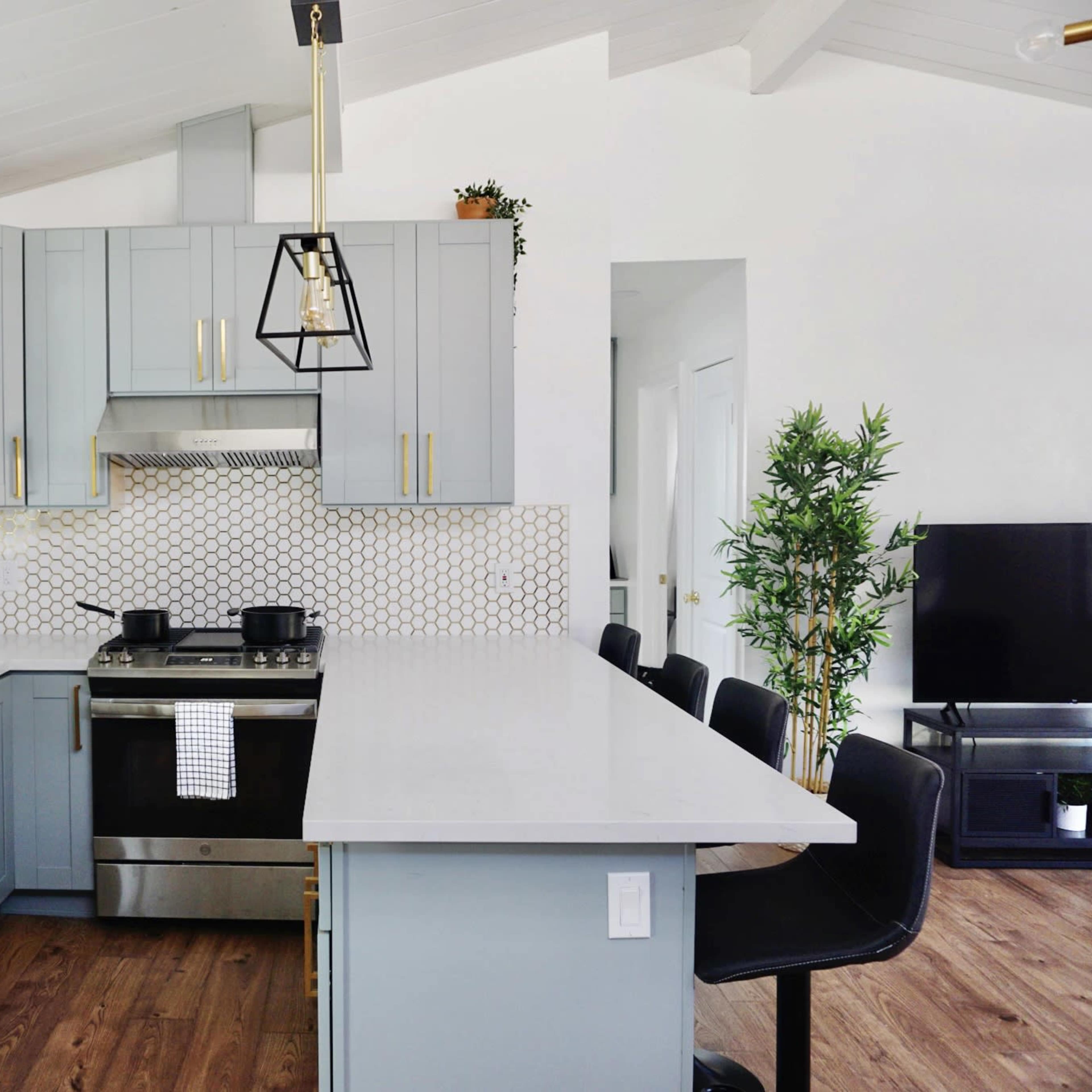 A modern kitchen with gray cabinets, a stainless steel stove, and white countertops, adjacent to a living area with a television.