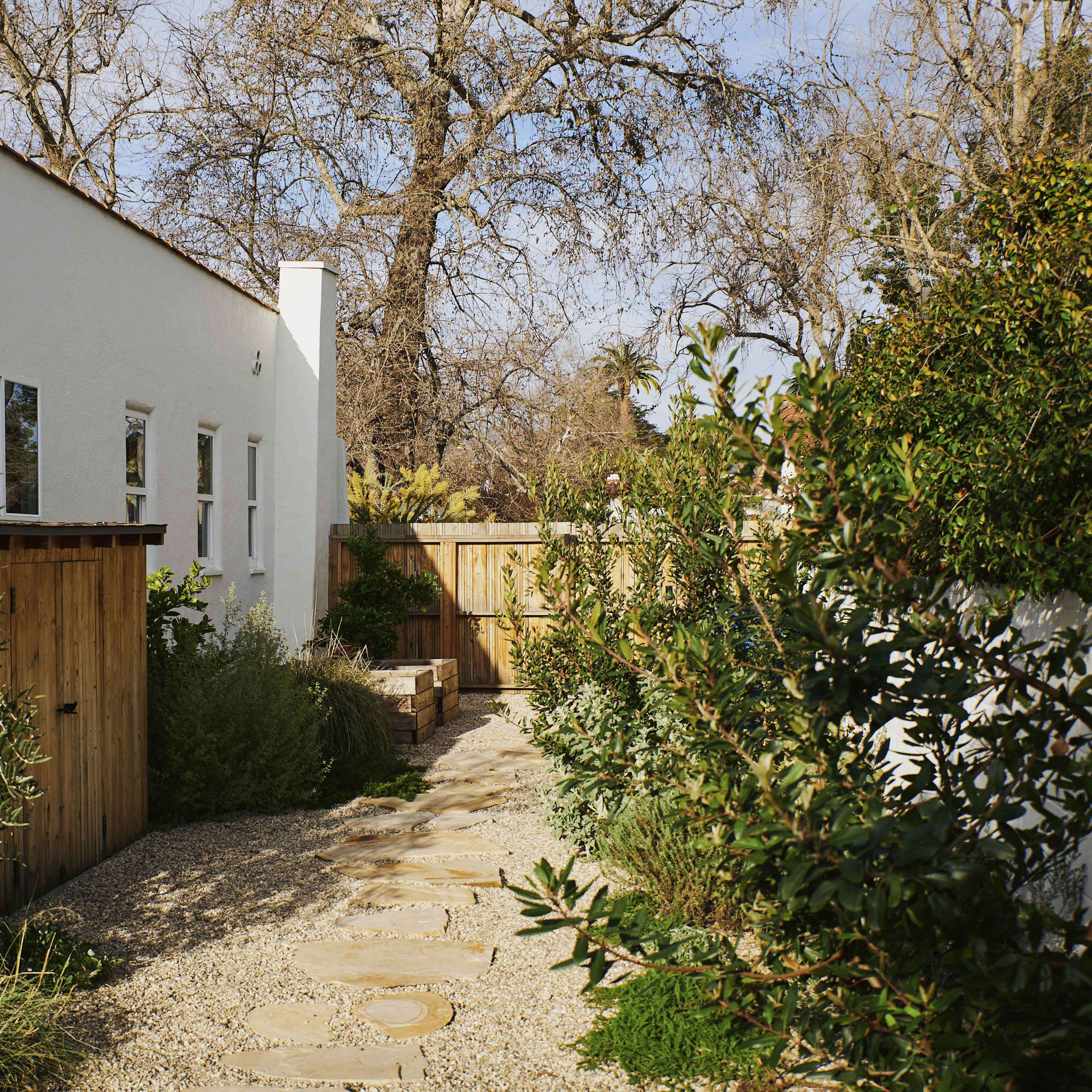 A stone path lined with greenery leads to a wooden gate beside a white building set in a tranquil outdoor space.