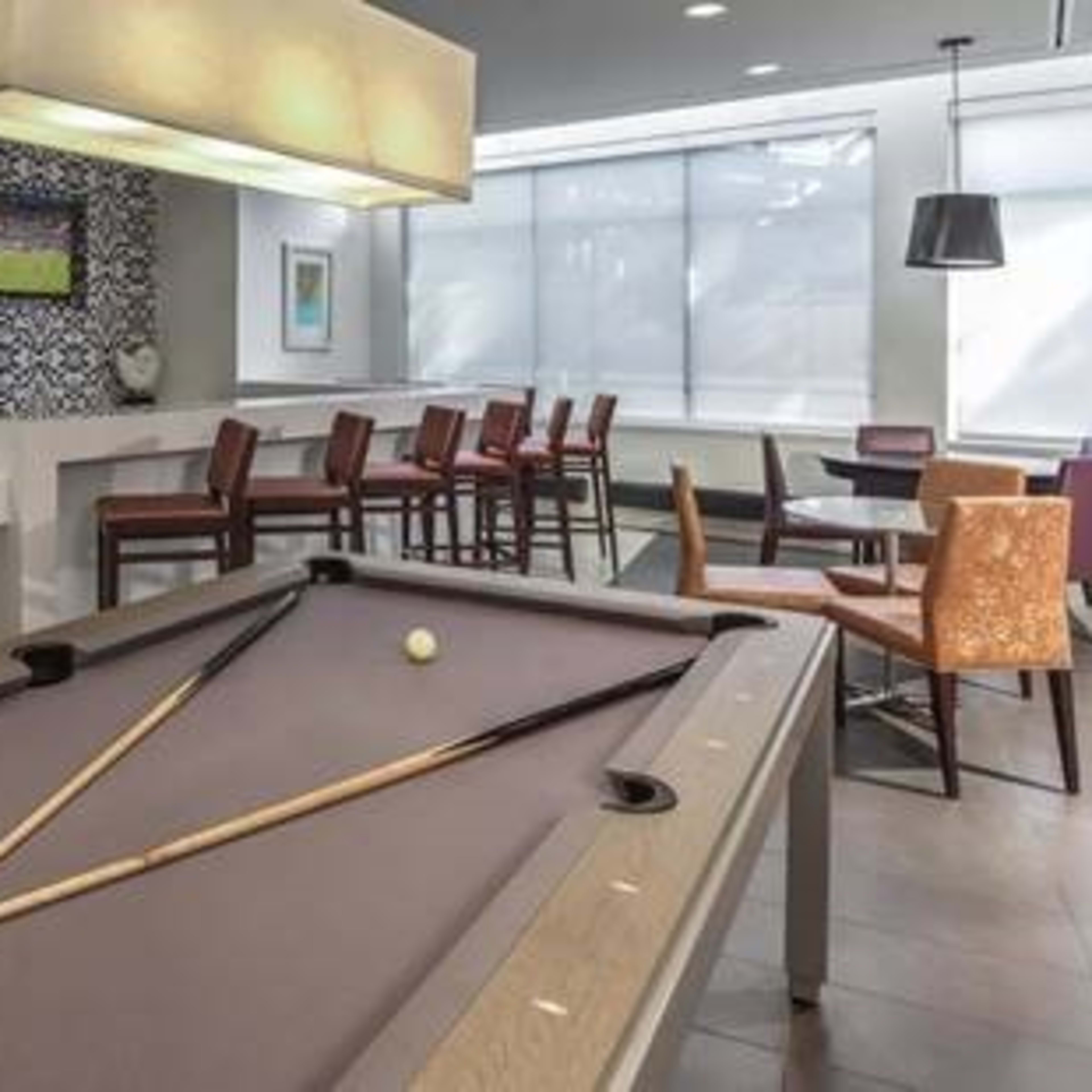 A modern recreational room featuring a pool table with a triangle rack of balls, surrounded by tables and chairs under bright lighting.