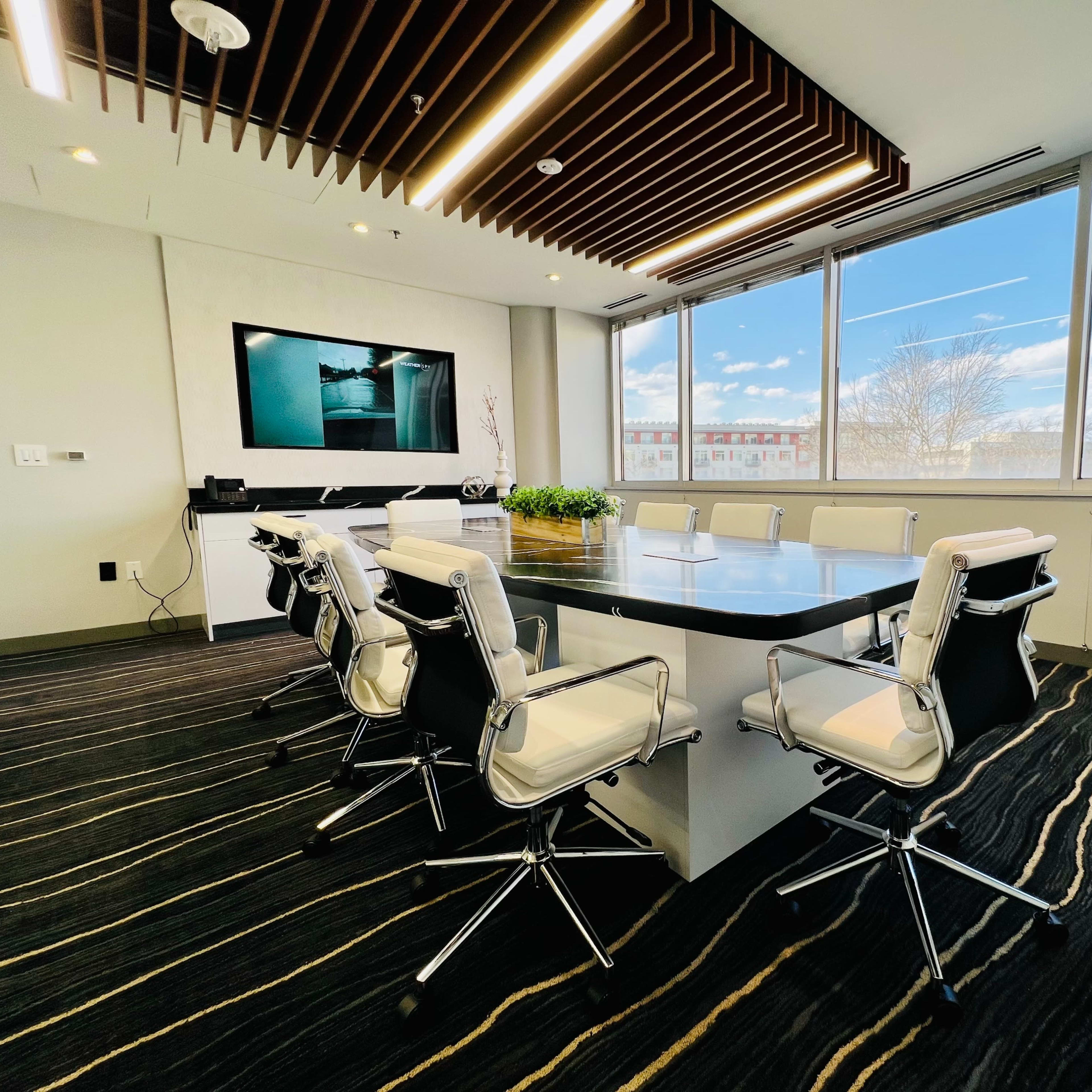 A modern conference room features a large table surrounded by white leather chairs, with a television mounted on the wall and large windows displaying a cloudy sky.