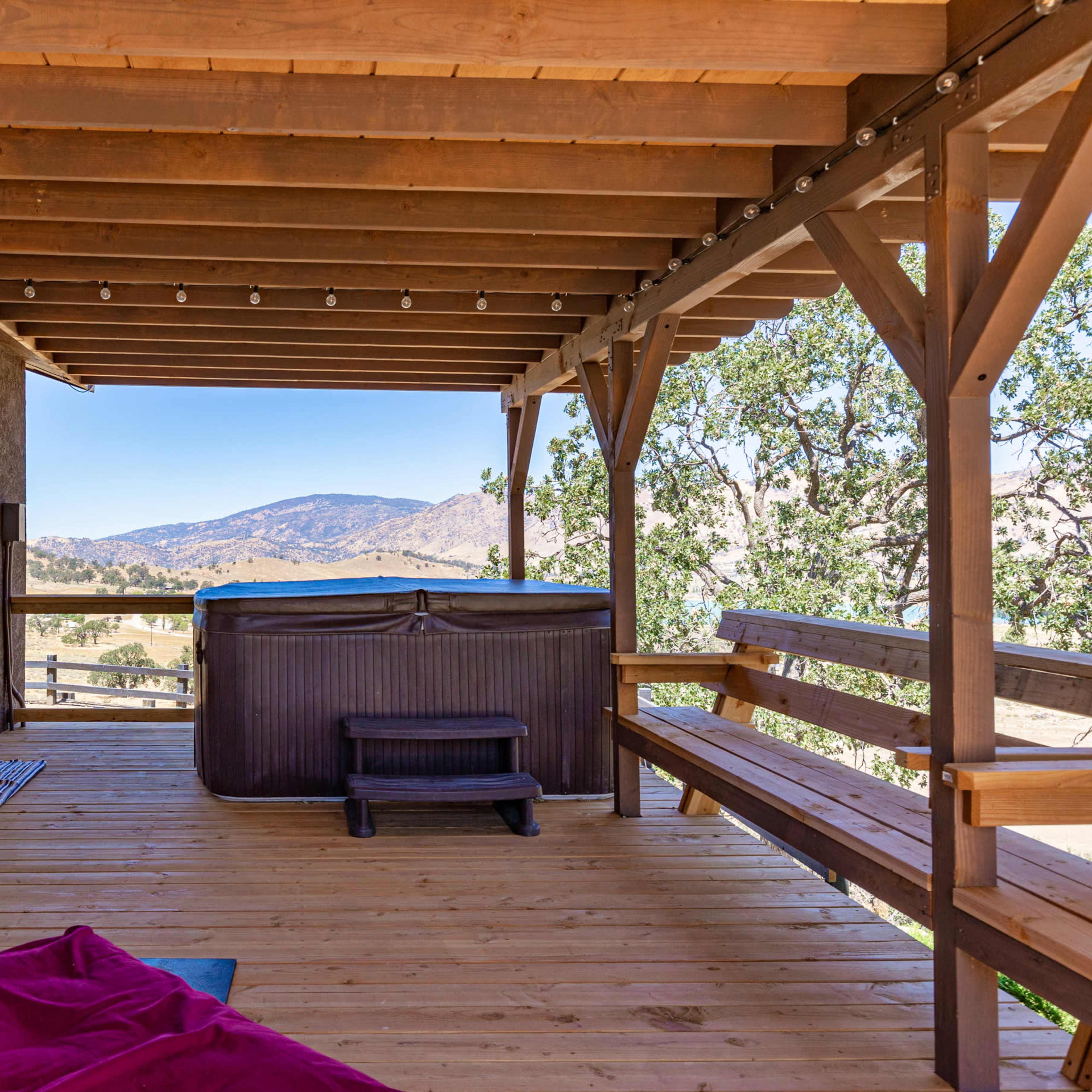 A wooden deck area featuring a hot tub, a grill, and seating, with a scenic view of mountains and trees in the background.