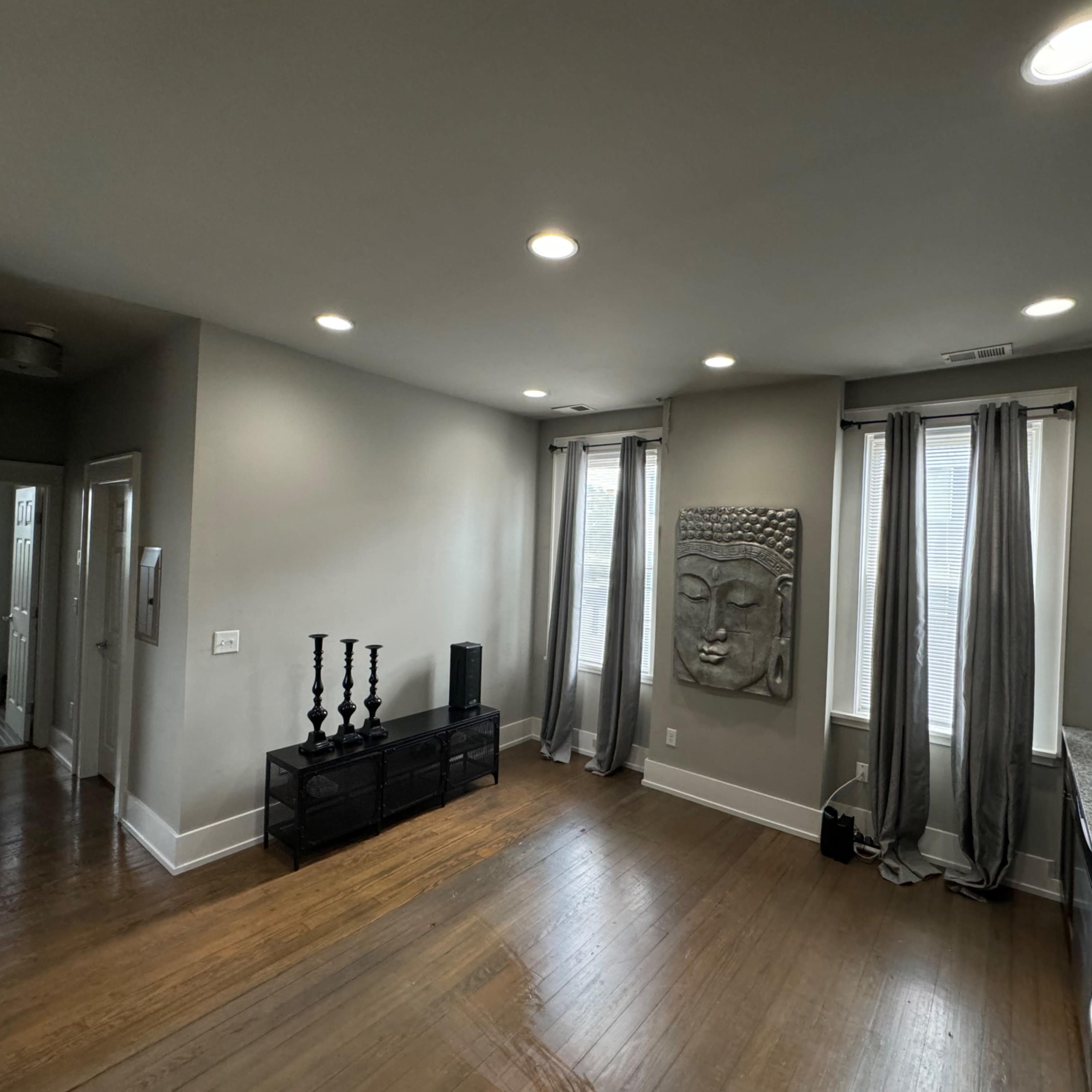 A contemporary living space featuring a textured wall sculpture, two tall windows with gray curtains, and a kitchen area with dark cabinets and a granite countertop.