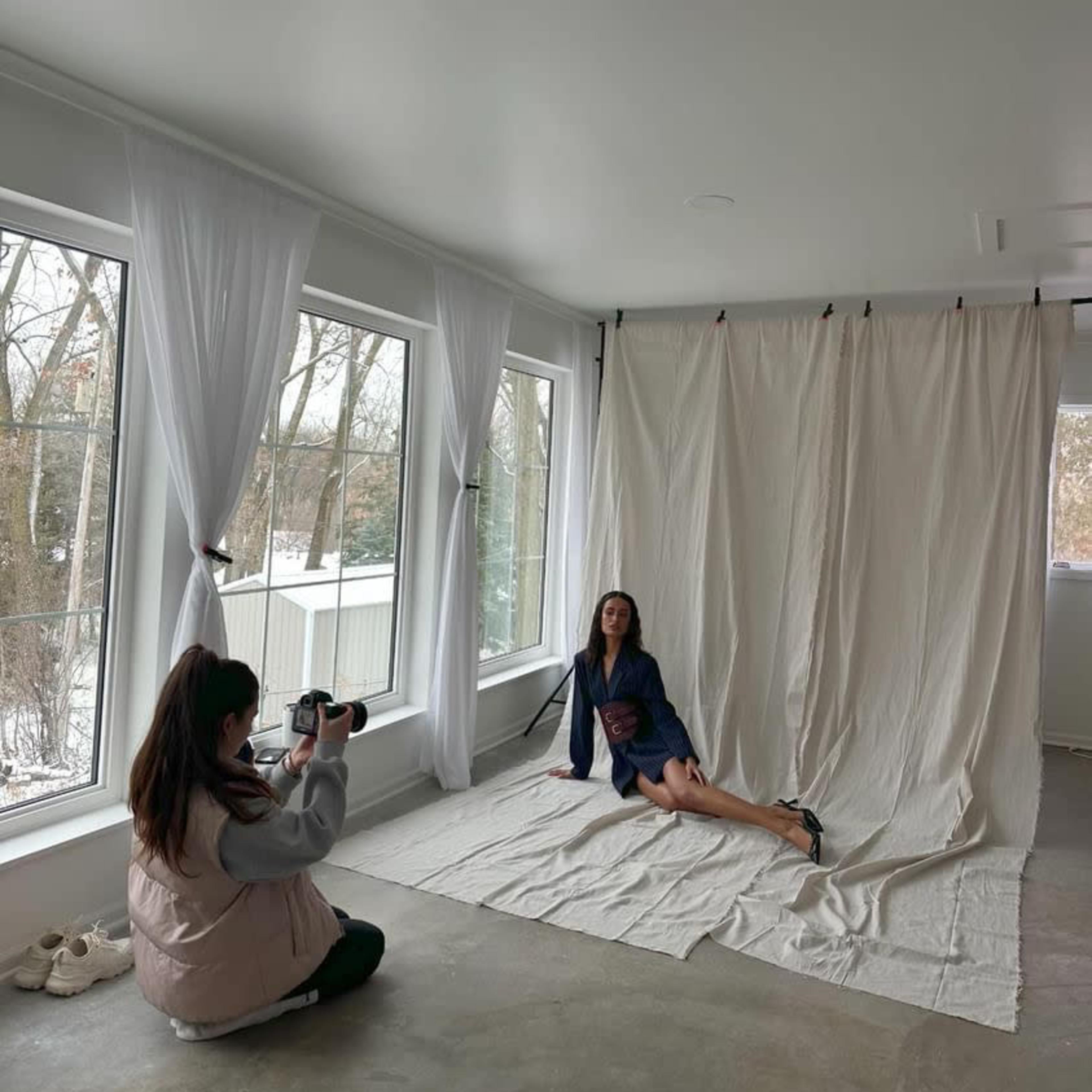 A photographer captures a model posing on a light-colored backdrop in a spacious room with large windows.