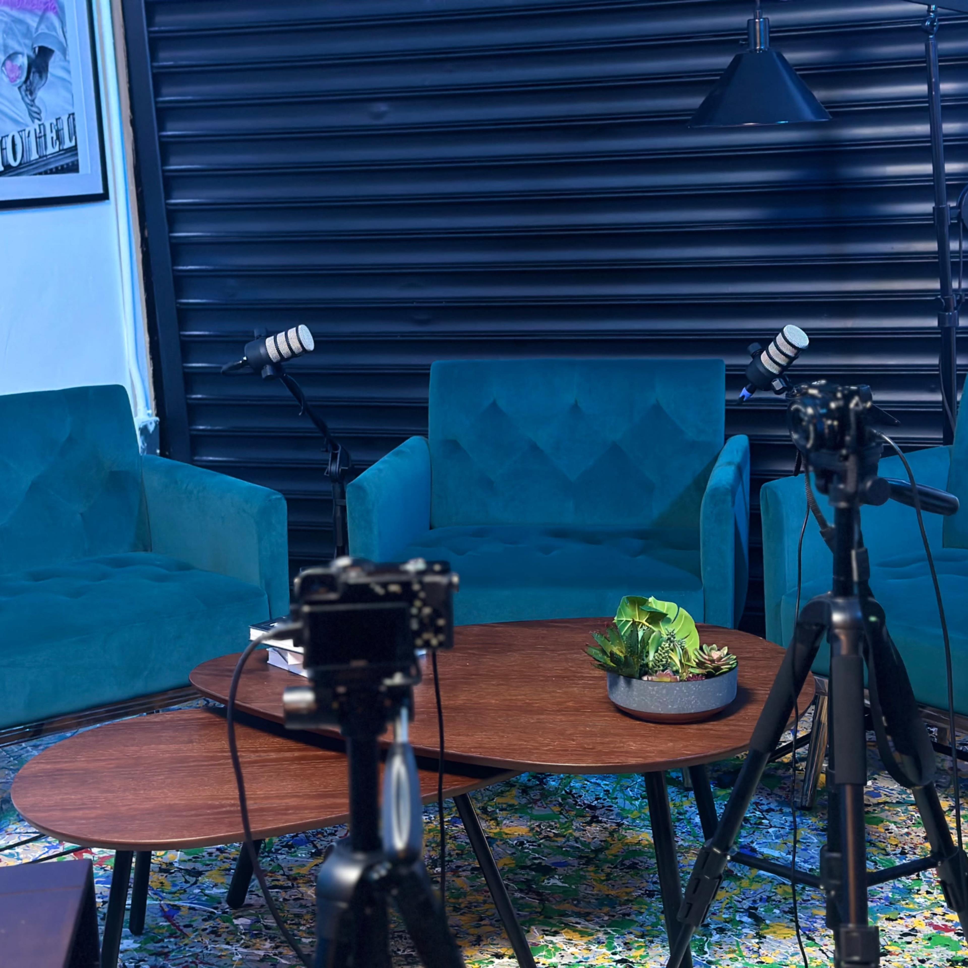 A cozy seating area with three teal chairs arranged around a wooden coffee table, accompanied by microphones and camera tripods, set against a dark slatted backdrop.