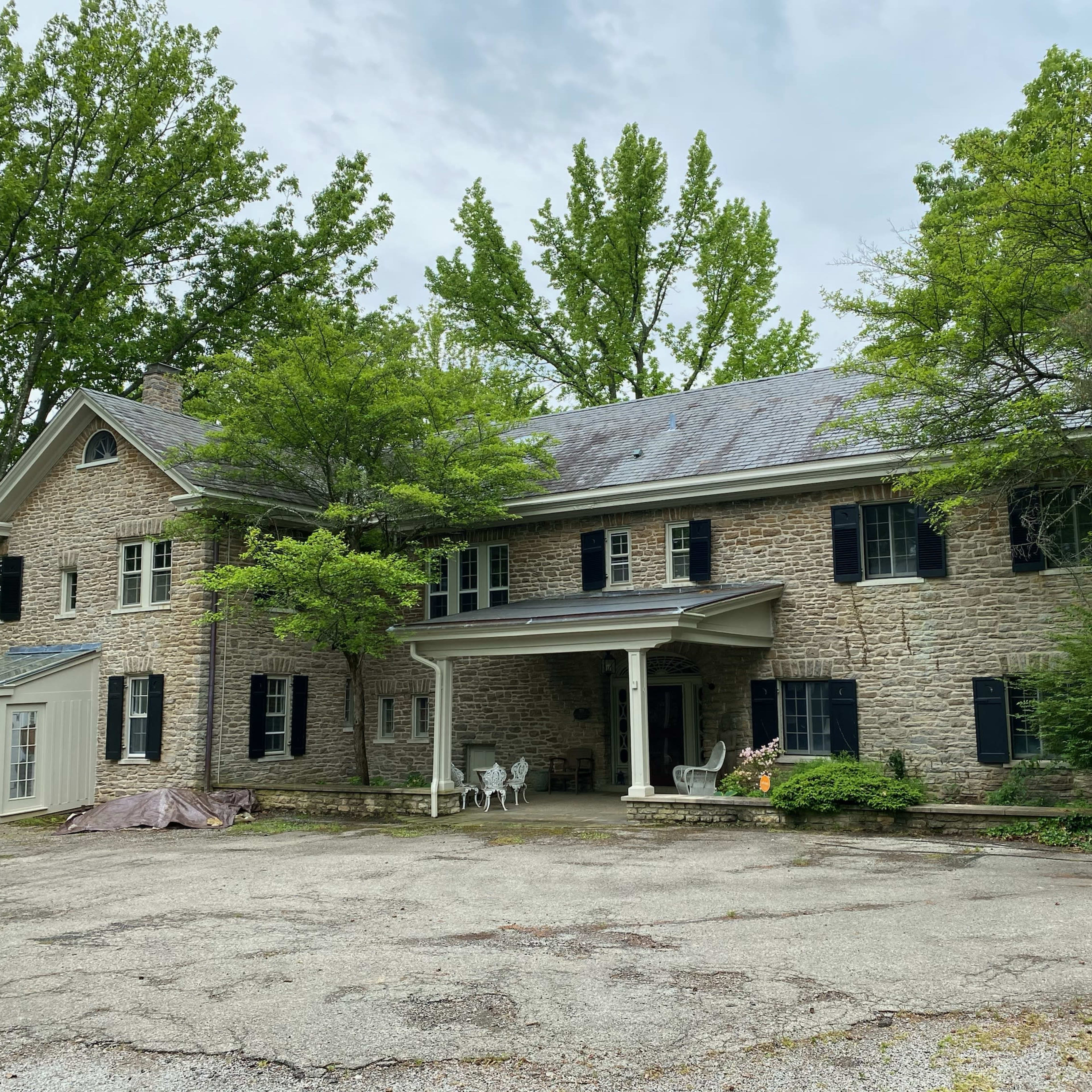 A large stone house surrounded by trees, with a gravel driveway and a covered porch in front.