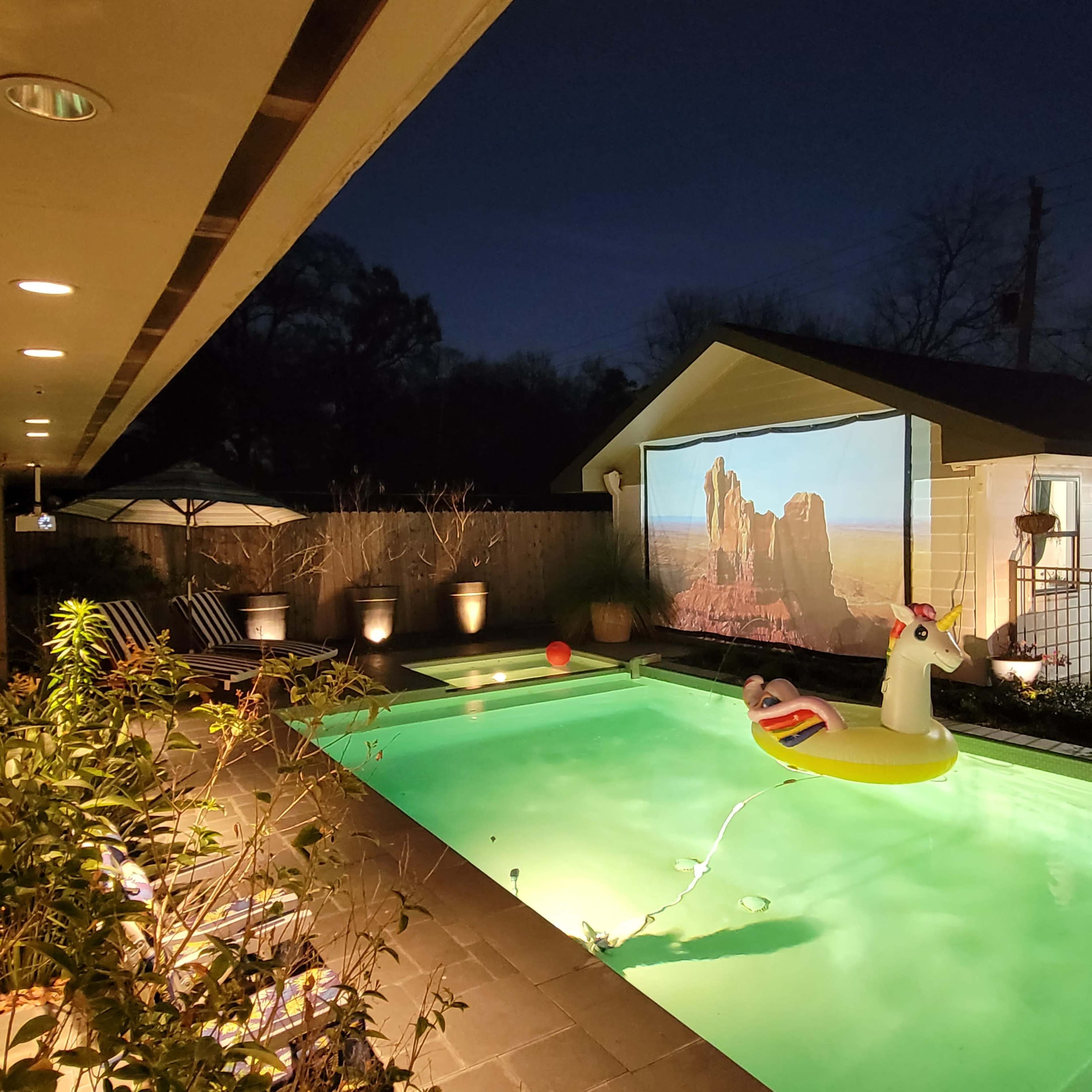 A pool area features a unicorn float, a movie screen showing a landscape, and illuminated outdoor plants under a night sky.
